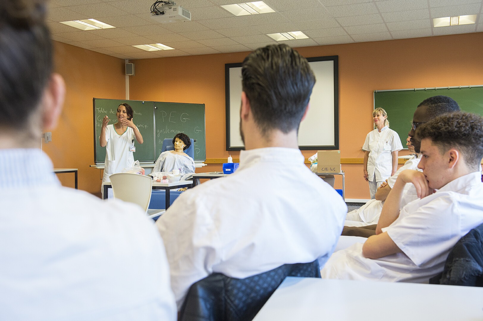 Un groupe de personnes en blouse blanche est assis et écoute une femme parler au premier plan d'une salle de classe. Un tableau noir, un écran et une autre femme debout près du tableau sont visibles, ainsi que les bureaux et le matériel de la classe.