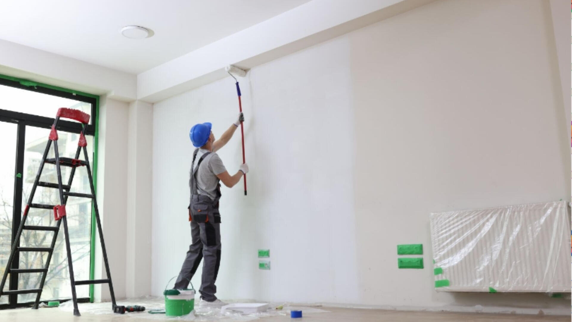 Une personne portant un casque bleu peint un mur blanc au rouleau dans une pièce lumineuse. Une échelle, un pot de peinture et un radiateur couvert se trouvent à proximité, tandis que du ruban adhésif vert délimite certaines parties du mur et du sol.