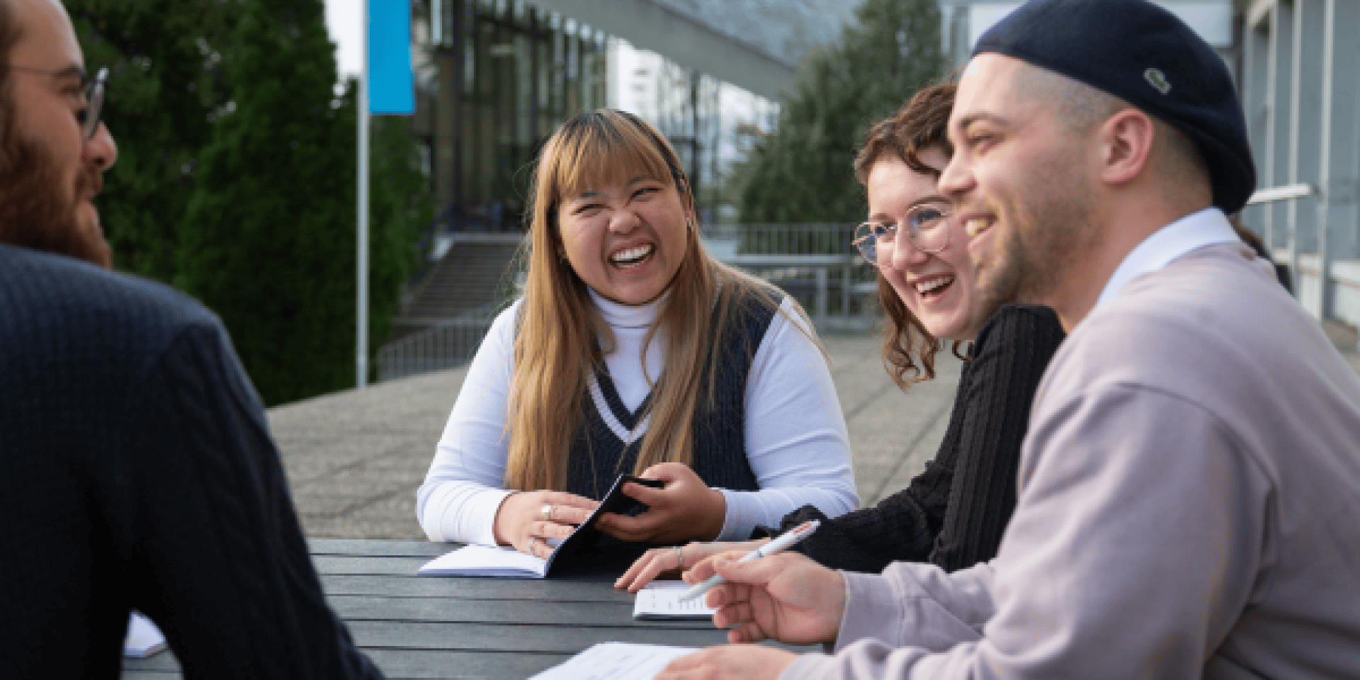 Quatre jeunes adultes sont assis à une table en plein air, souriant et riant ensemble tout en tenant des papiers et des stylos, semblant étudier ou travailler sur un projet de groupe. Des arbres et un bâtiment sont visibles à l'arrière-plan.