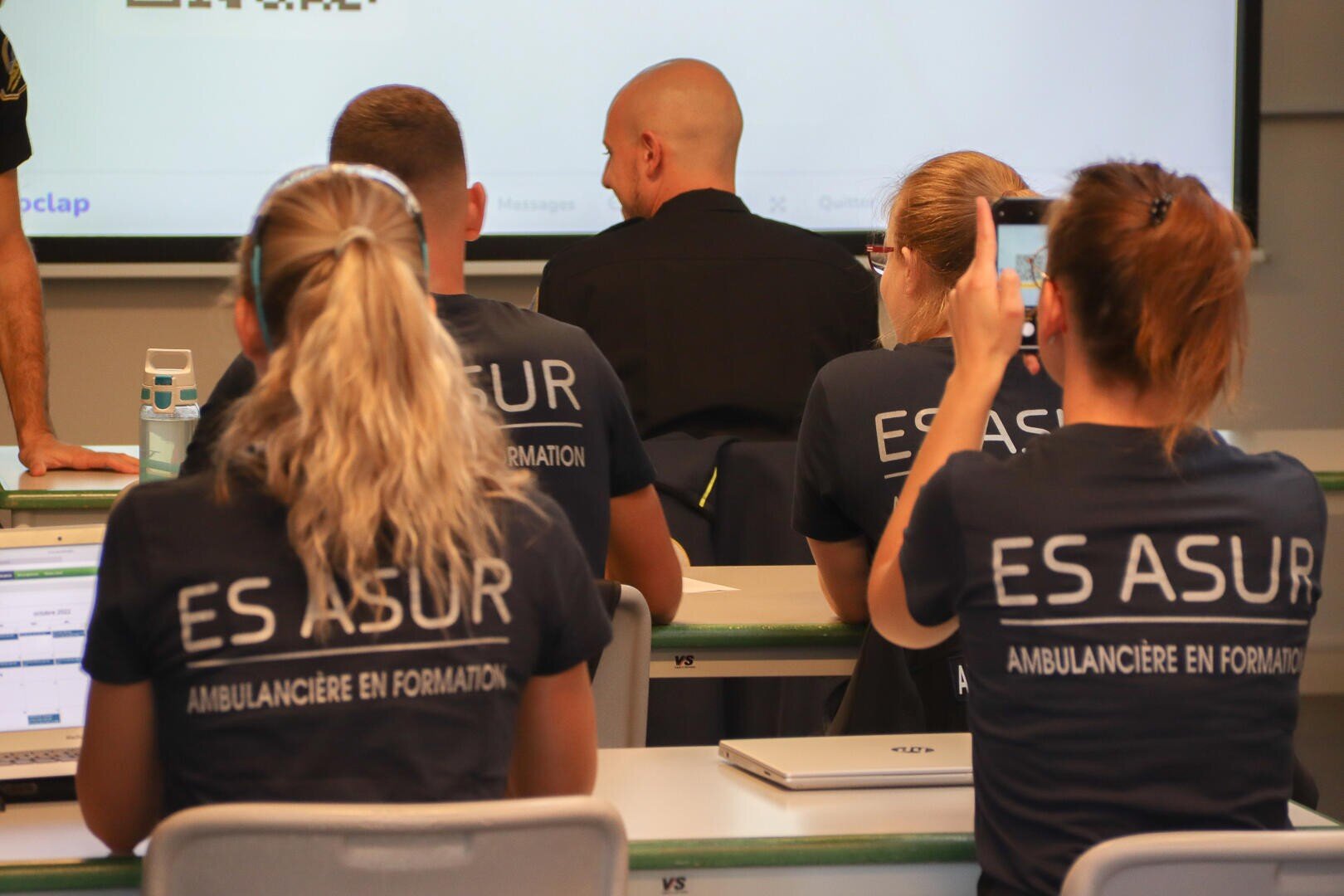 Plusieurs personnes sont assises dans une salle de classe, vue de dos. Elles portent des chemises bleu marine avec "ES ASUR AMBULANCIÈRE EN FORMATION" imprimé dans le dos. Une personne brandit un téléphone pour enregistrer ou prendre une photo.