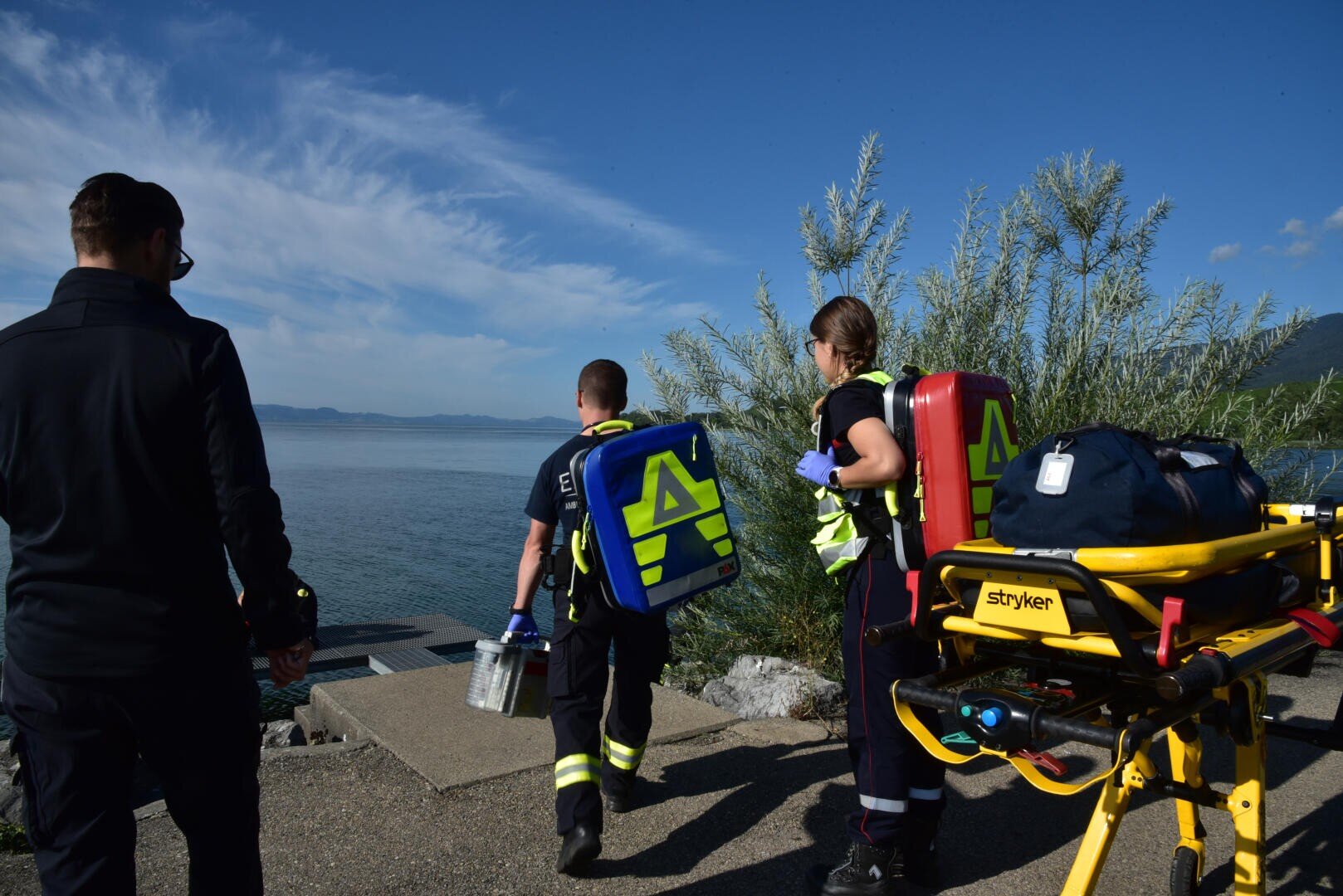 Trois ambulanciers en uniforme se dirigent vers un lac en transportant du matériel médical. Une civière contenant du matériel se trouve sur la droite. La scène se déroule par une journée claire et ensoleillée.