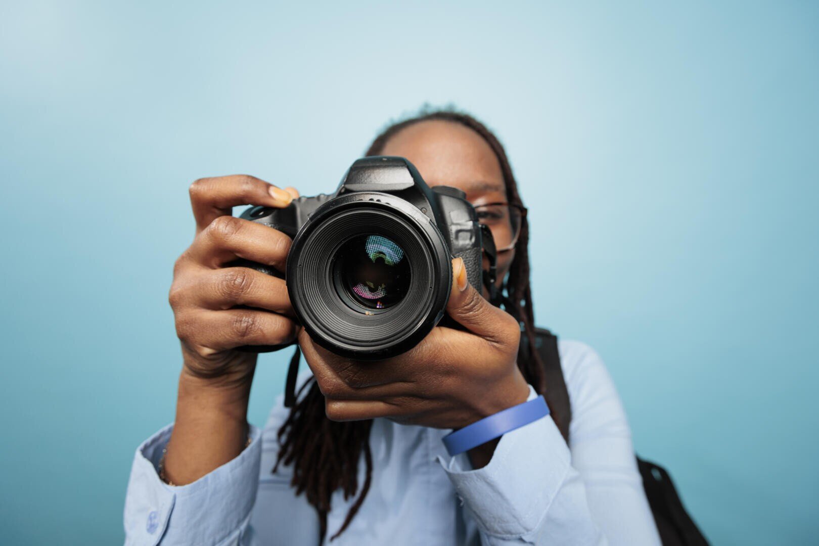 Une personne aux cheveux longs, portant des lunettes et une chemise bleue, tient un appareil photo reflex numérique à hauteur de son visage, en orientant l'objectif vers le spectateur, sur un fond bleu clair uni.