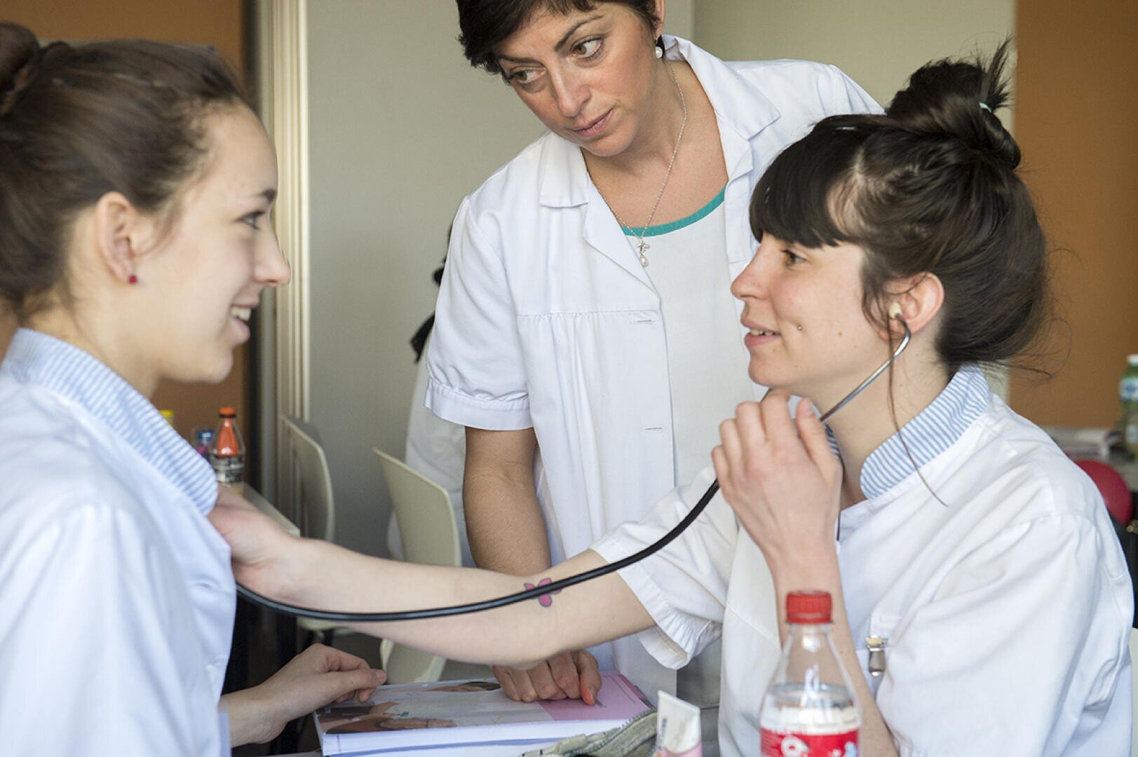 Trois femmes en blouse blanche ; l'une utilise un stéthoscope pour écouter la poitrine de l'autre tandis que la troisième l'observe. Elles semblent se trouver dans un environnement clinique ou dans une salle de classe.