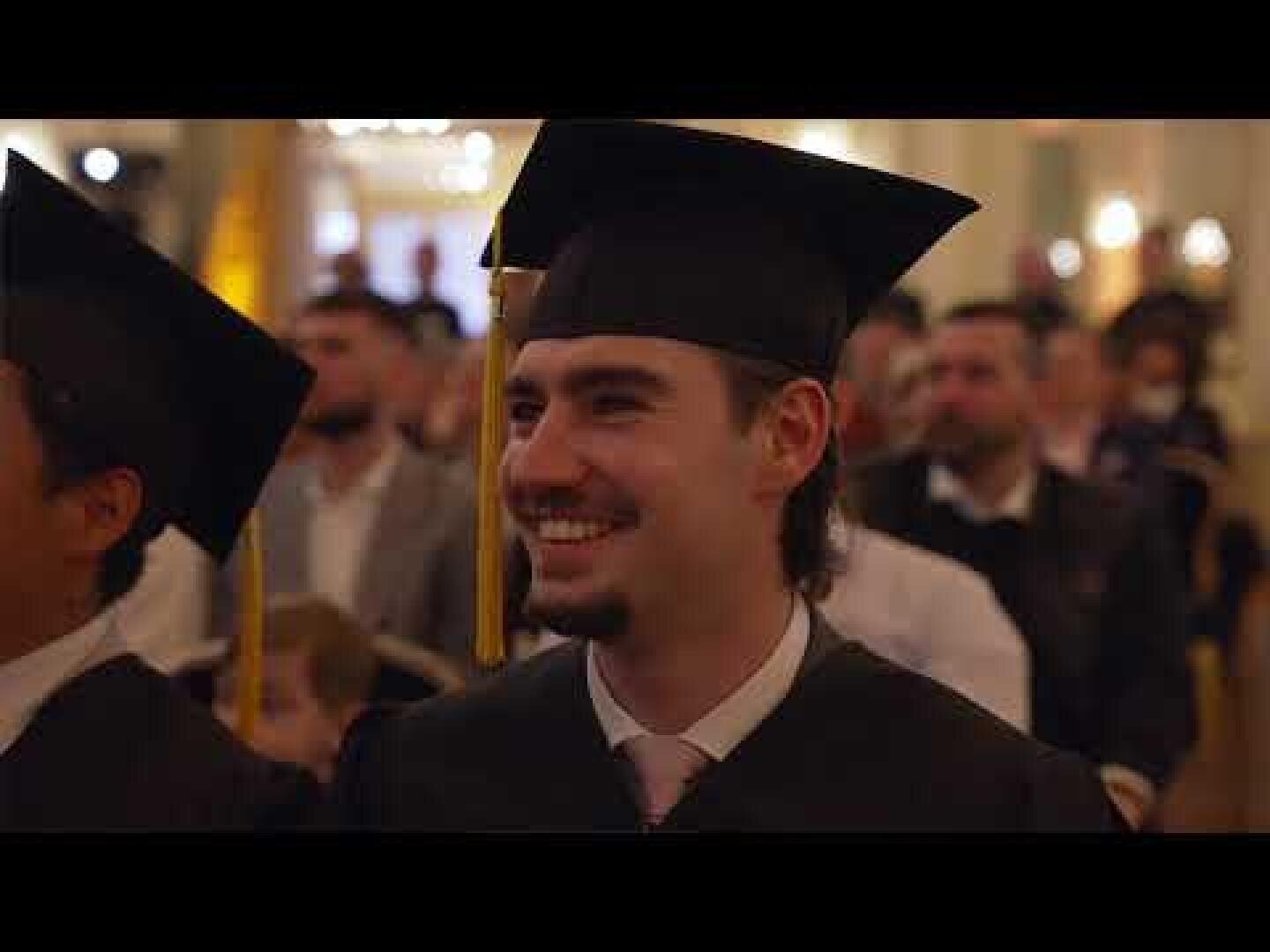 Un jeune homme, coiffé d'une toque et vêtu d'une toge, sourit, assis parmi d'autres diplômés lors d'une cérémonie. À l'arrière-plan, on aperçoit le public dans une salle baignée d'une lumière chaleureuse.