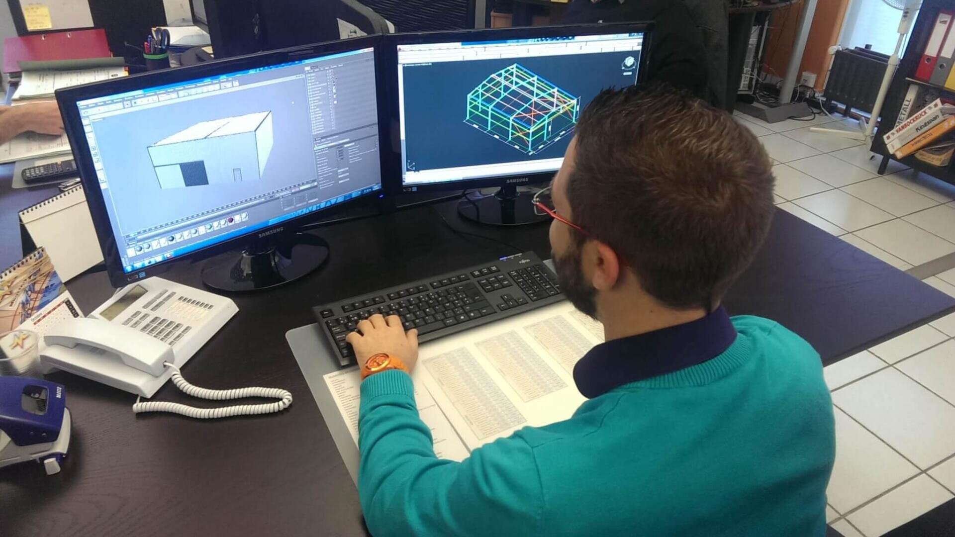 Une personne travaille à un bureau où se trouvent deux écrans d'ordinateur affichant des modèles architecturaux en 3D. Elle utilise un clavier et une souris, des documents et un téléphone sont posés sur le bureau.