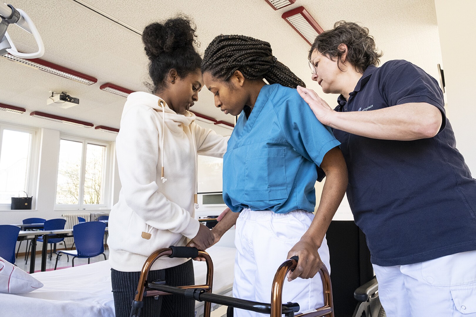 Deux femmes assistent une personne en tenue médicale et utilisant un déambulateur dans une salle clinique lumineuse, probablement pour une rééducation physique ou une thérapie. Une femme la soutient de côté, tandis que l'autre la stabilise de face.