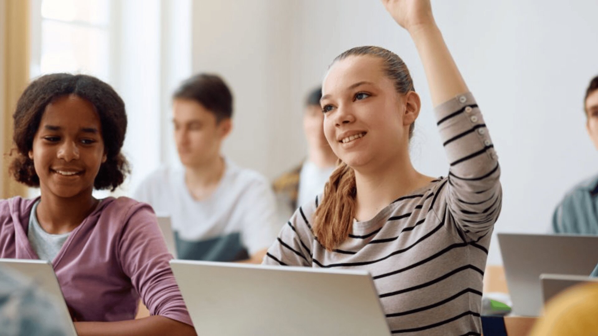 Une élève souriante lève la main dans une salle de classe, assise à côté d'un camarade. Les deux élèves ont des ordinateurs portables ouverts sur leurs bureaux, tandis que d'autres élèves travaillent en arrière-plan.