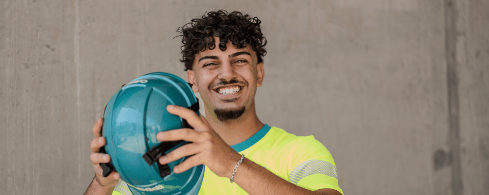 Un jeune homme souriant aux cheveux bouclés tient un casque de sécurité sarcelle devant lui, vêtu d'une chemise jaune fluo, debout contre un simple mur de béton.