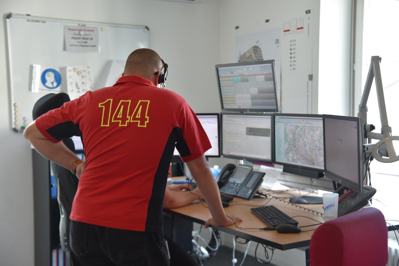 Une personne portant une chemise rouge avec le numéro 144 dans le dos se tient à un bureau avec plusieurs écrans d'ordinateur, dans ce qui semble être un centre d'appel d'urgence ou un bureau de répartition.