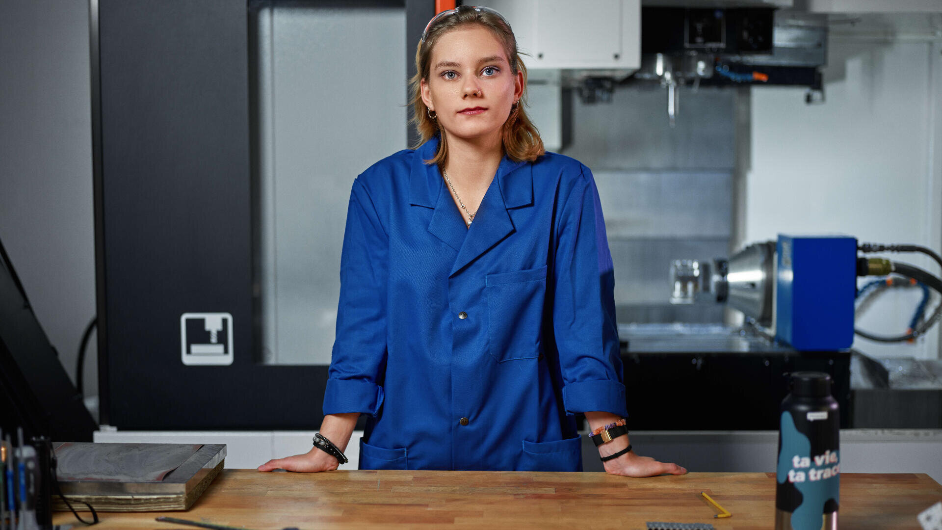 Une personne portant une blouse bleue se tient derrière un établi en bois dans un atelier ou un laboratoire, avec des machines et divers outils visibles à l'arrière-plan.