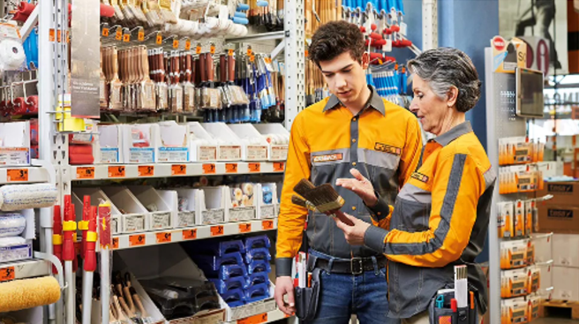 Deux employés de quincaillerie en uniformes orange et gris discutent des pinceaux devant un étalage de fournitures de peinture, notamment des pinceaux, des rouleaux et des plateaux.
