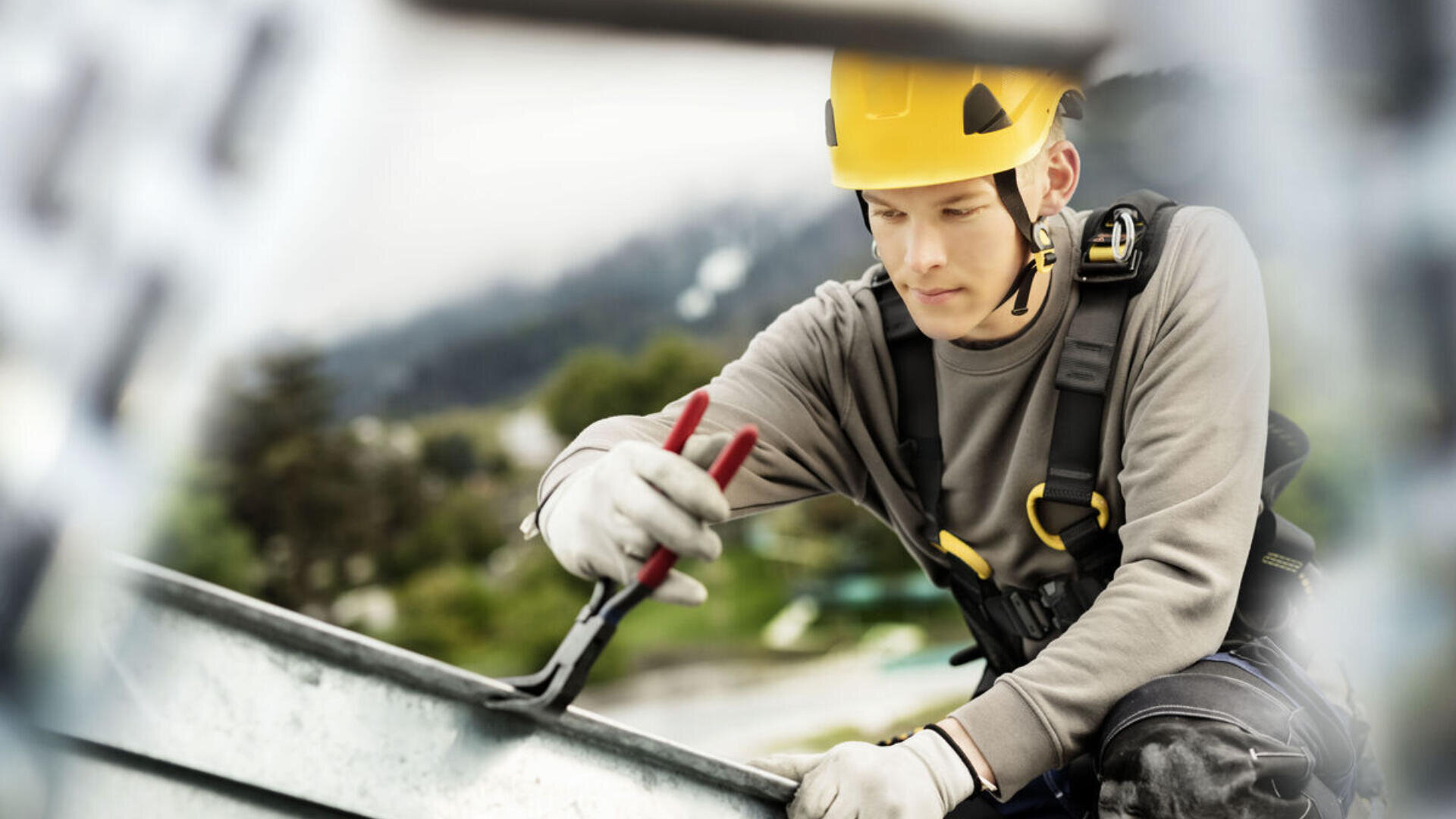 Un ouvrier de la construction portant un casque jaune et un harnais de sécurité utilise des pinces tout en travaillant sur un toit métallique à l'extérieur. Des arbres et un arrière-plan flou indiquent un lieu élevé.