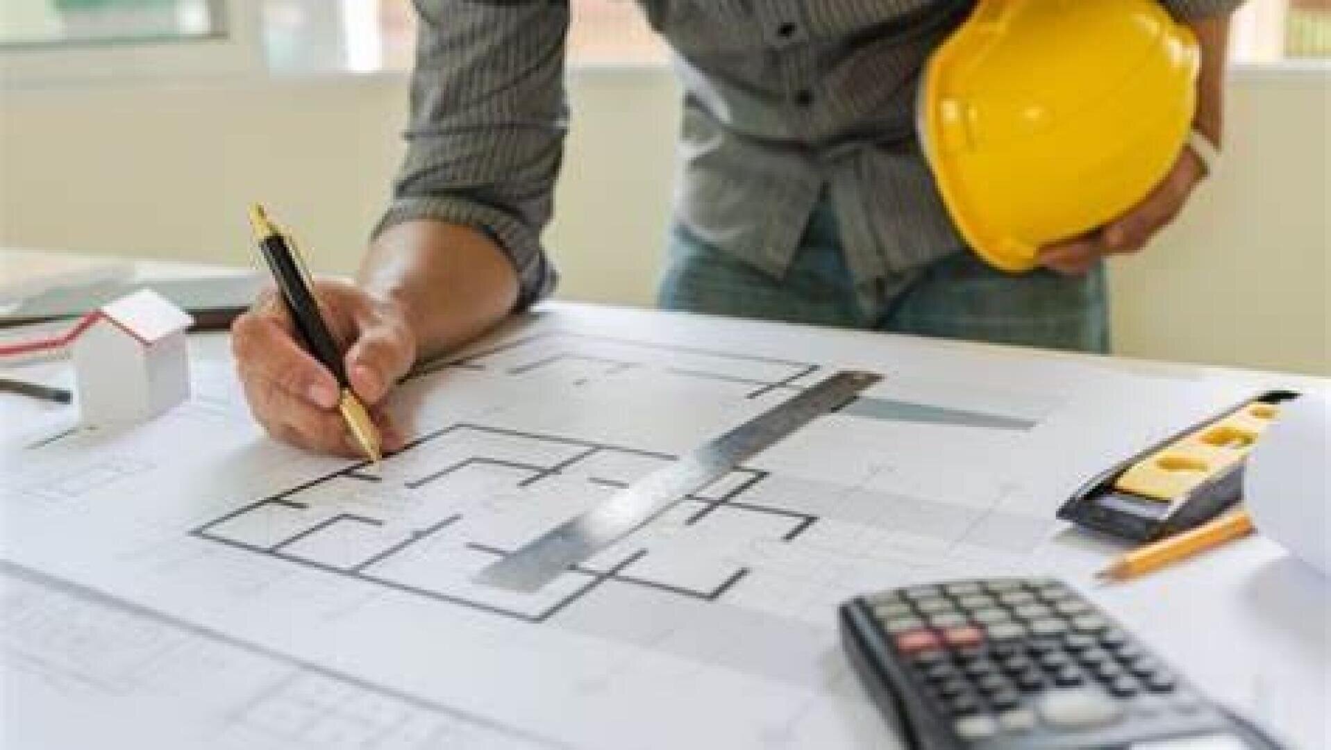 Une personne portant un casque jaune examine des plans d'architecture sur une table, à l'aide d'un crayon et d'une règle. À proximité se trouvent une calculatrice, un mètre ruban et une petite maquette de maison.