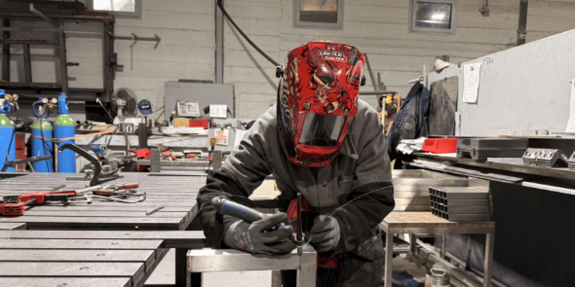 Une personne portant un casque de soudage rouge et un équipement de protection soude du métal sur un établi dans un atelier industriel, avec divers outils et équipements visibles à l'arrière-plan.