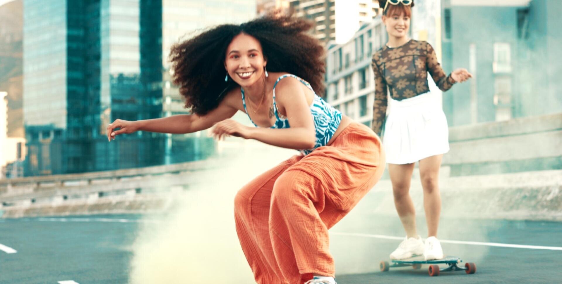 Deux jeunes femmes font du skateboard dans une rue de la ville. La femme à l'avant a les cheveux bouclés, un pantalon orange et un débardeur à motifs, et sourit en faisant du skate. La femme derrière porte un haut à fleurs transparent et une jupe blanche, avec de la fumée colorée en arrière-plan.