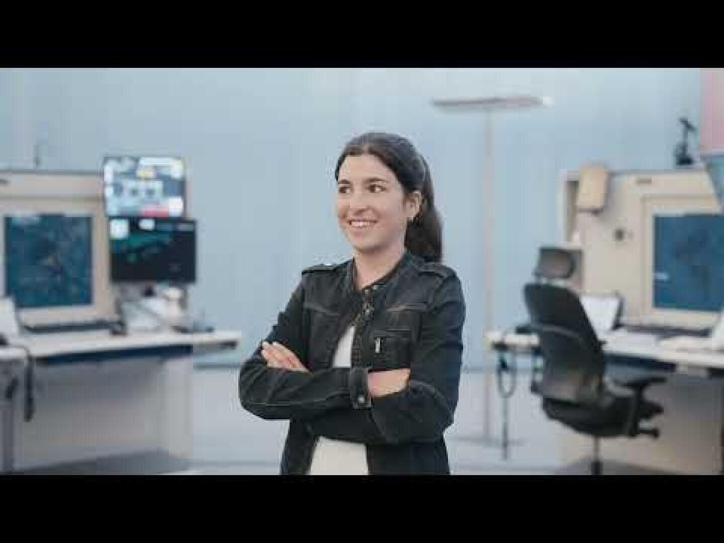 Une femme aux cheveux noirs, vêtue d'une veste noire et d'une chemise blanche, sourit, les bras croisés, dans une salle de contrôle moderne avec des écrans d'ordinateur et des chaises à l'arrière-plan.