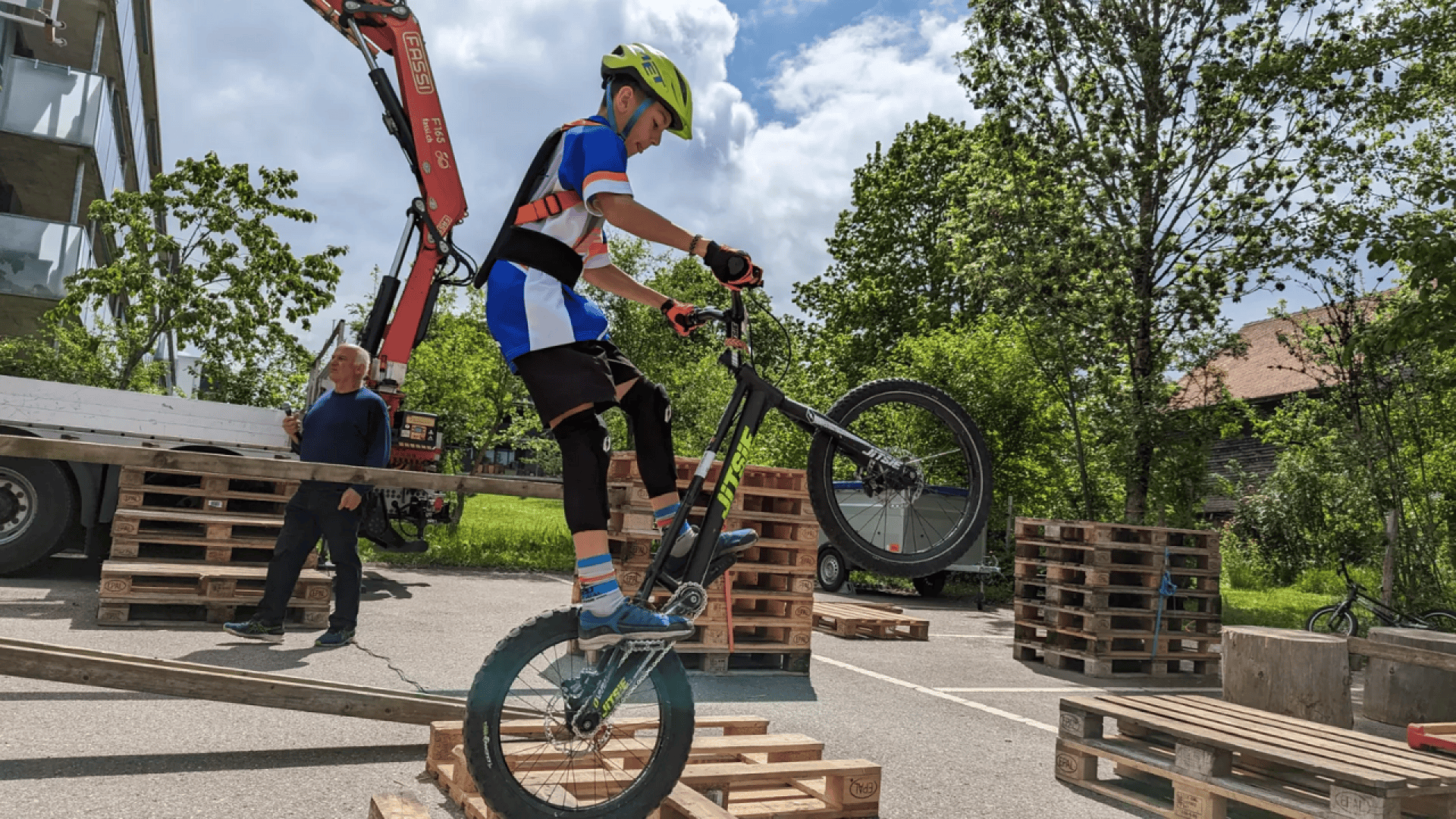 Un enfant portant un casque et un équipement de protection fait du vélo sur des palettes en bois empilées, en soulevant la roue avant, tandis qu'un adulte se tient à proximité, en plein air, par une journée ensoleillée.