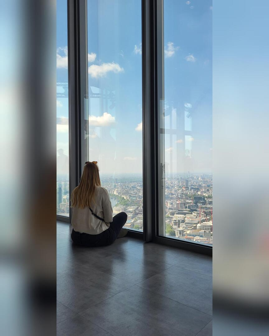 Une femme aux longs cheveux blonds est assise sur le sol, les jambes croisées, près de grandes baies vitrées, regardant la ligne d'horizon d'une ville par une journée claire et ensoleillée.