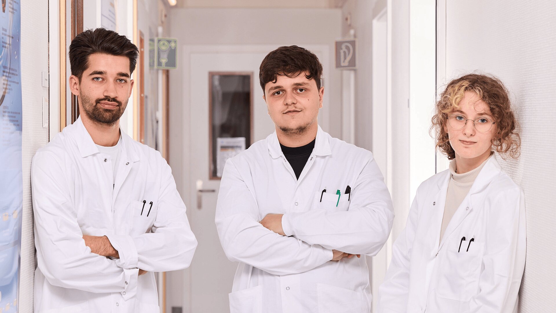 Trois jeunes scientifiques portant des blouses blanches se tiennent côte à côte dans un couloir, face à la caméra, avec des expressions neutres et les bras croisés. Des affiches et des portes de laboratoire sont visibles à l'arrière-plan.