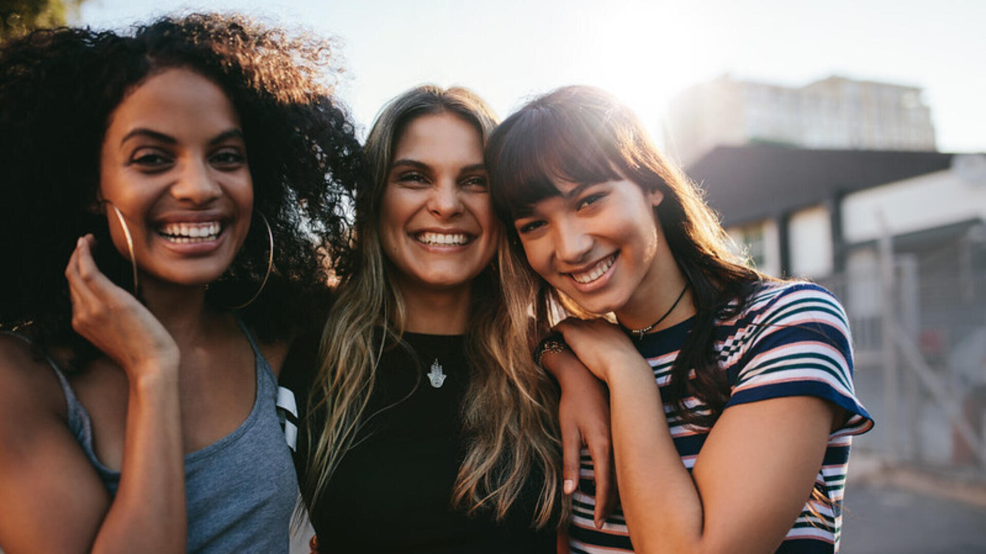 Trois jeunes femmes se tiennent côte à côte en plein air et sourient à l'appareil photo. Elles se tiennent par le bras, rayonnant d'amitié et de bonheur, avec un arrière-plan urbain ensoleillé derrière elles.