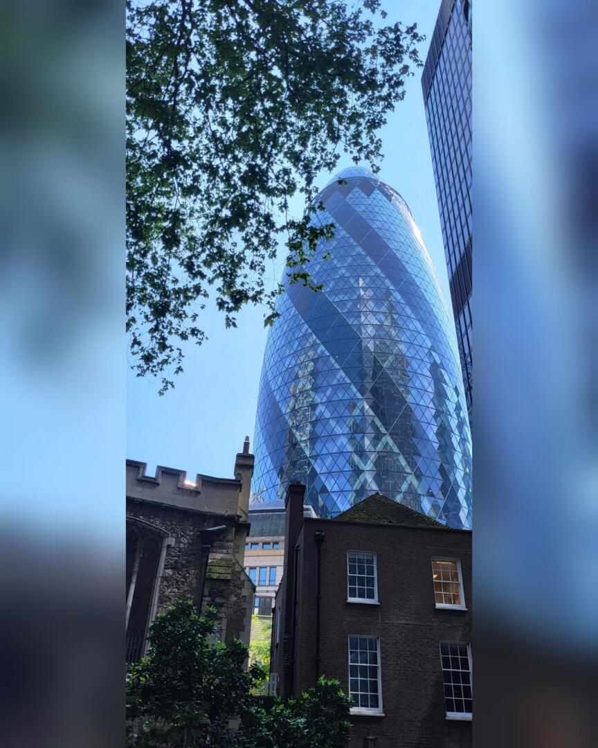 Le gratte-ciel Gherkin, à Londres, s'élève derrière des bâtiments en briques plus anciens et des branches d'arbres feuillus, sous un ciel bleu limpide.