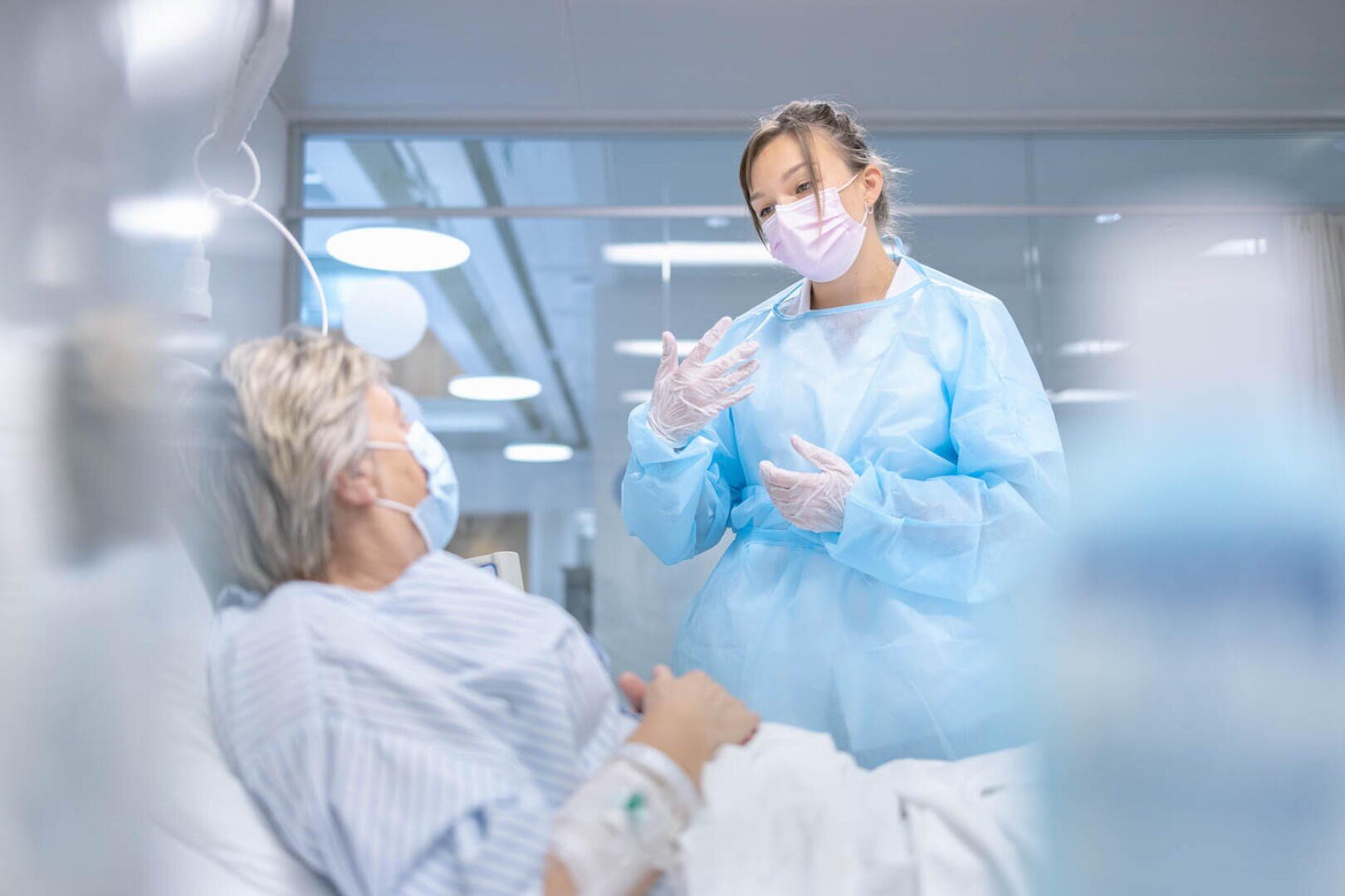 Un agent de santé en tenue de protection complète et portant un masque se tient à côté d'un lit d'hôpital, s'adressant à un patient allongé et portant lui aussi un masque. Le décor semble être une chambre d'hôpital moderne.