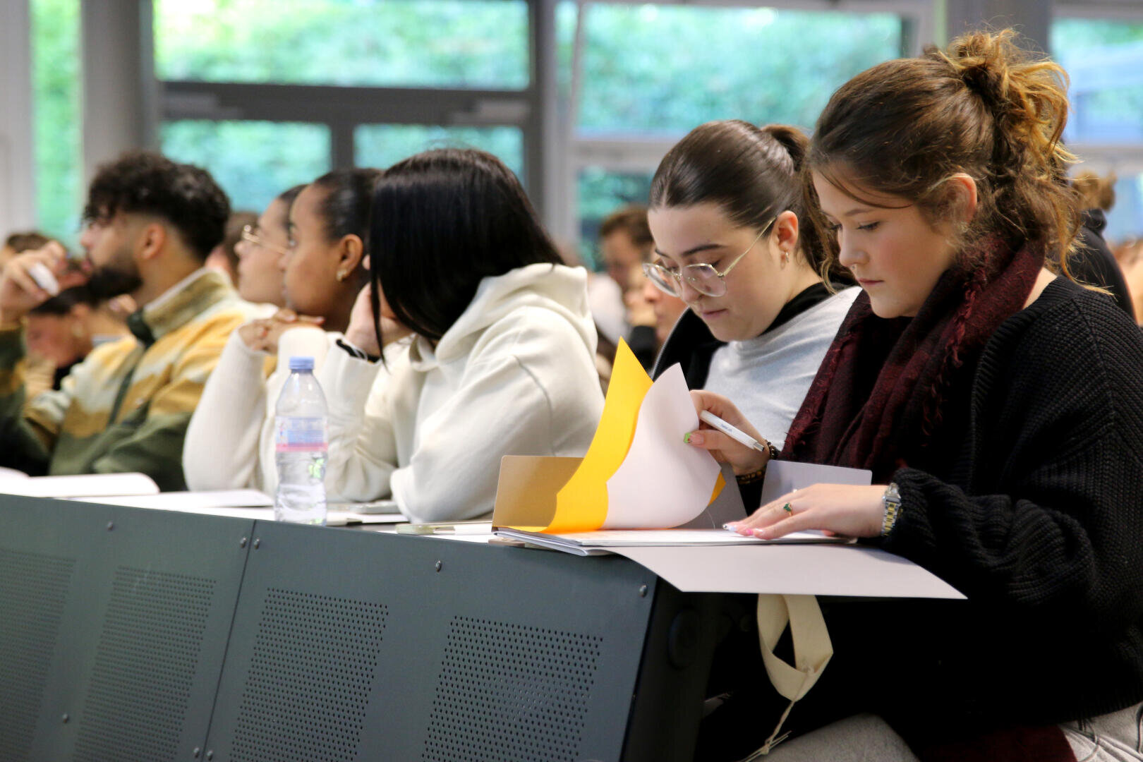 Les élèves sont assis dans une salle de classe ; certains prennent des notes, d'autres écoutent attentivement. Un élève feuillette des feuilles, tandis qu'un autre lit. Des bouteilles d'eau et des cahiers sont posés sur les pupitres. De grandes fenêtres donnent sur la verdure à l'extérieur.