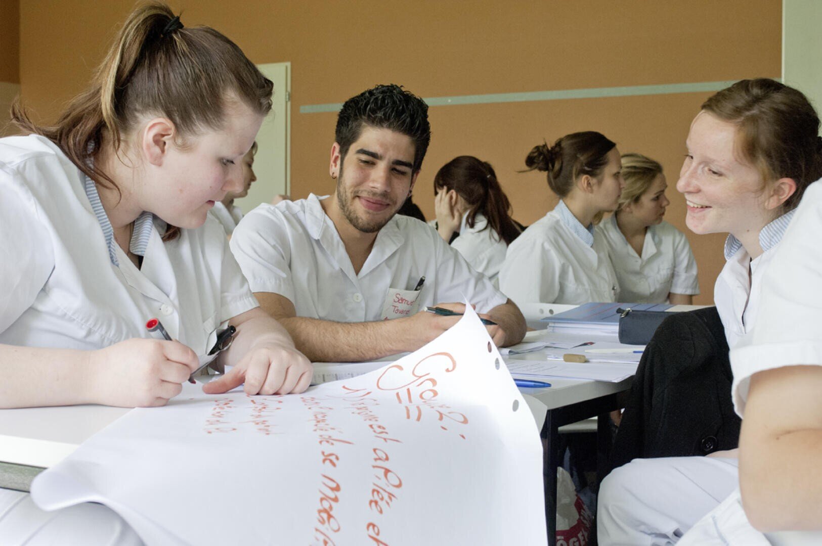 Un groupe d'étudiants en soins infirmiers en uniforme blanc sont assis à une table, souriant et travaillant sur une grande feuille de papier au cours d'une activité en classe. D'autres étudiants sont visibles en train de travailler à l'arrière-plan.