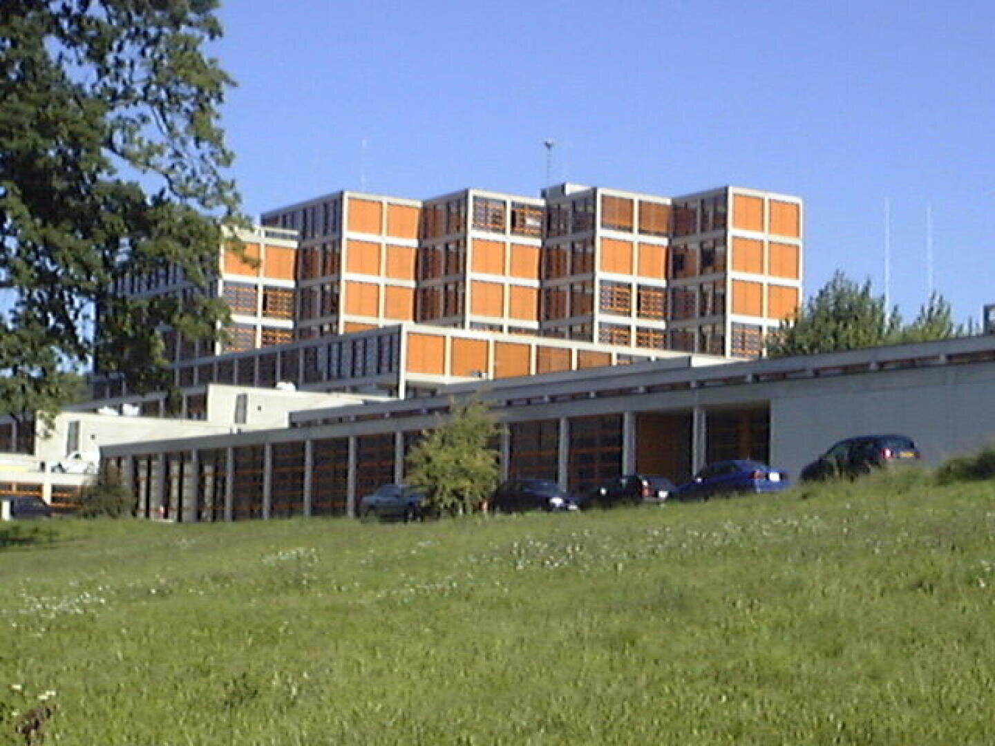 Un bâtiment de plusieurs étages avec une façade orange et blanche en forme de grille se dresse derrière une structure plus basse et des voitures garées, entouré d'herbe verte et d'arbres sous un ciel bleu clair.