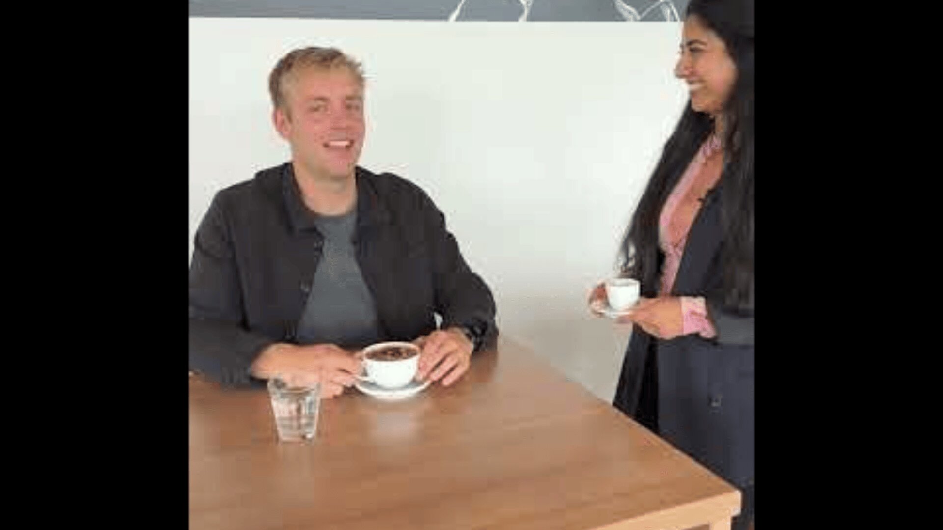 Un homme est assis à une table en bois, souriant, avec une tasse de café devant lui, tandis qu'une femme se tient à côté de la table, tenant une petite tasse et lui souriant.