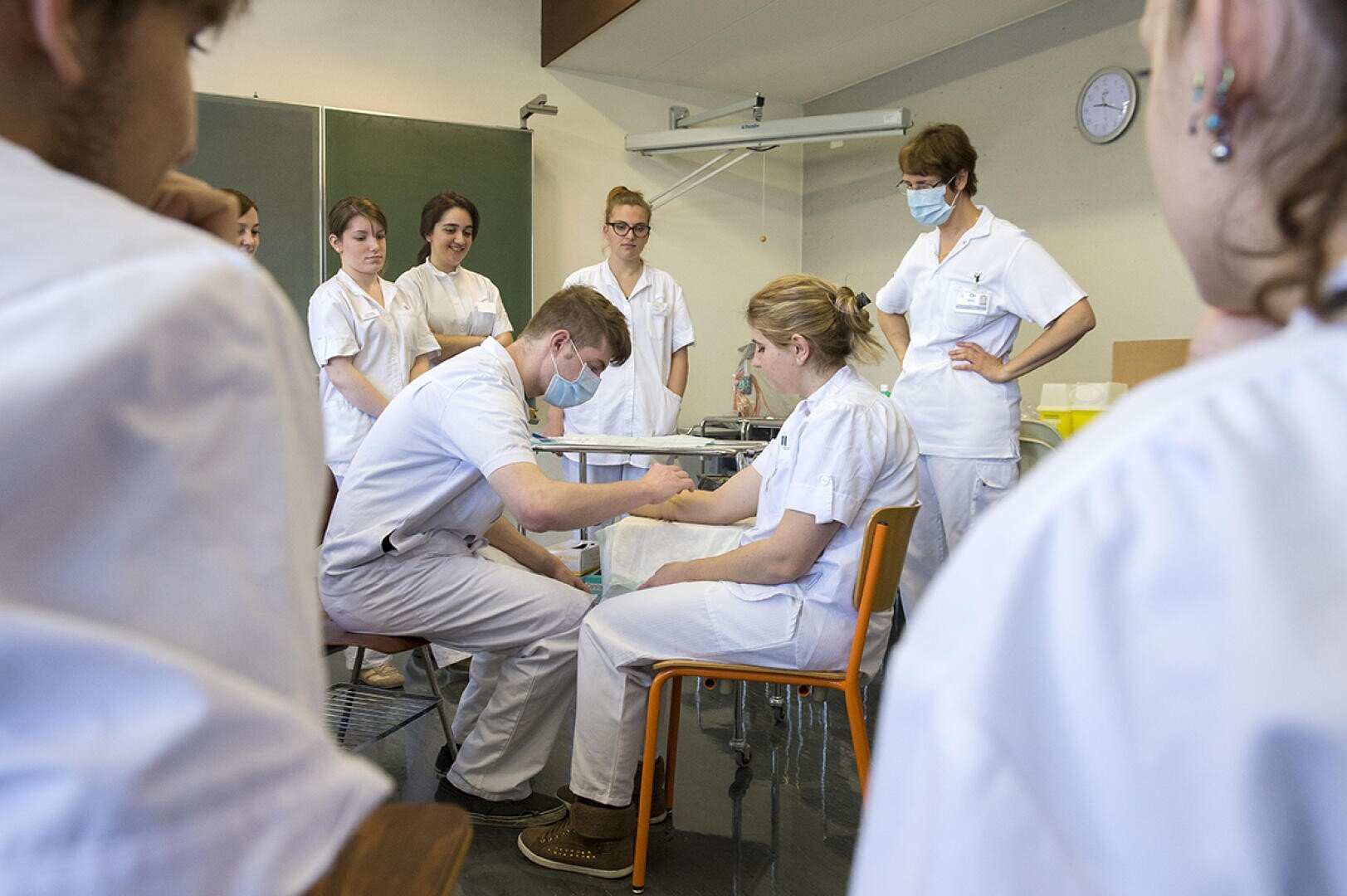 Un groupe d'étudiants en médecine en uniforme blanc pratique une procédure médicale dans une salle de classe. Un étudiant, portant un masque, panse le bras d'un autre étudiant tandis que les autres observent attentivement.