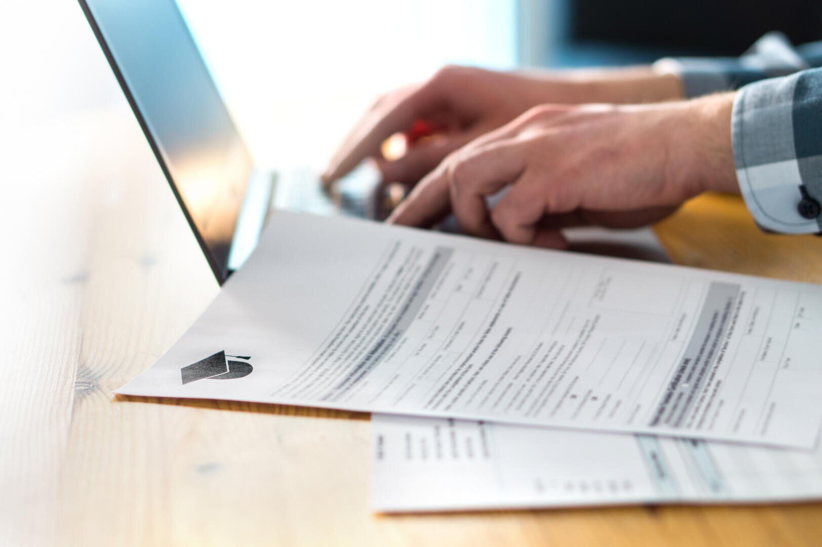 Une personne tapant sur un ordinateur portable, avec des documents, dont un formulaire comportant du texte et un trombone, posés sur un bureau en bois au premier plan. La scène évoque un travail mêlant tâches administratives et tâches numériques.