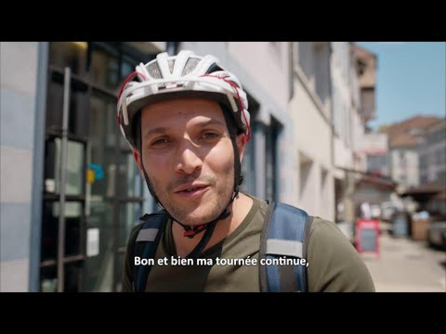 Un homme portant un casque de vélo blanc et un sac à dos se tient dans une rue de la ville, regardant la caméra. Les sous-titres français de l'image indiquent : "Bon et bien ma tournée continue".