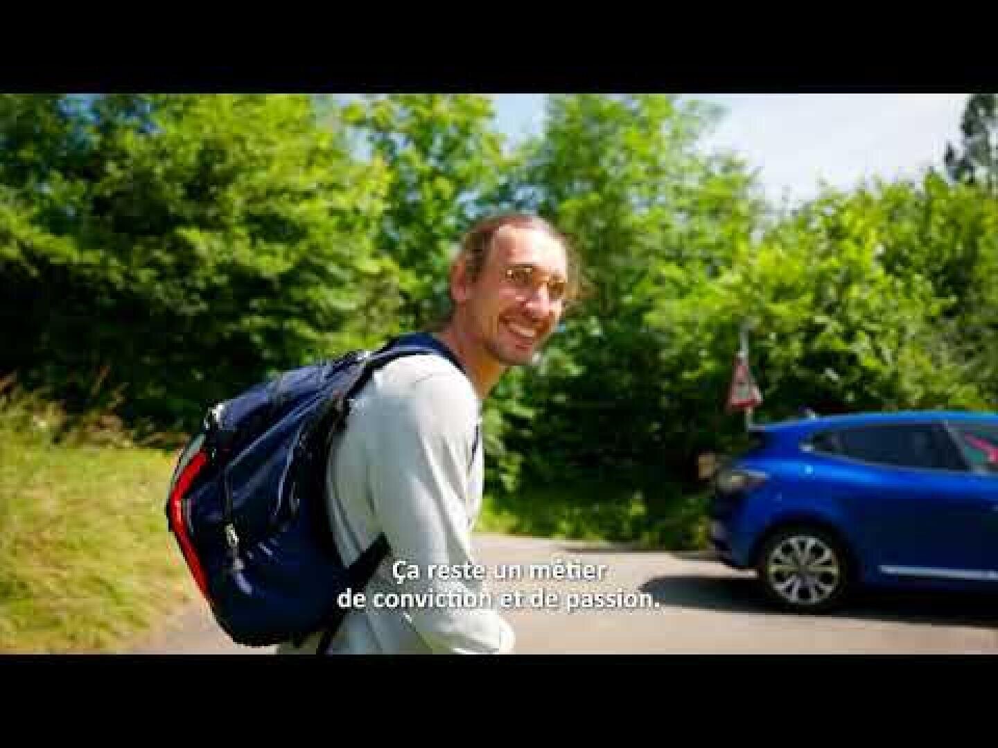 Un homme portant un sac à dos sourit à la caméra alors qu'il se tient à l'extérieur près d'une voiture bleue, avec des arbres verts en arrière-plan. Des sous-titres en français apparaissent au bas de l'image.