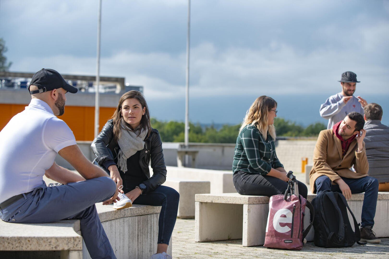 Un groupe de jeunes adultes est assis par deux sur des bancs extérieurs, discutant et se détendant avec des sacs à dos à proximité. Le ciel est nuageux et l'arrière-plan montre de la verdure et des bâtiments modernes.