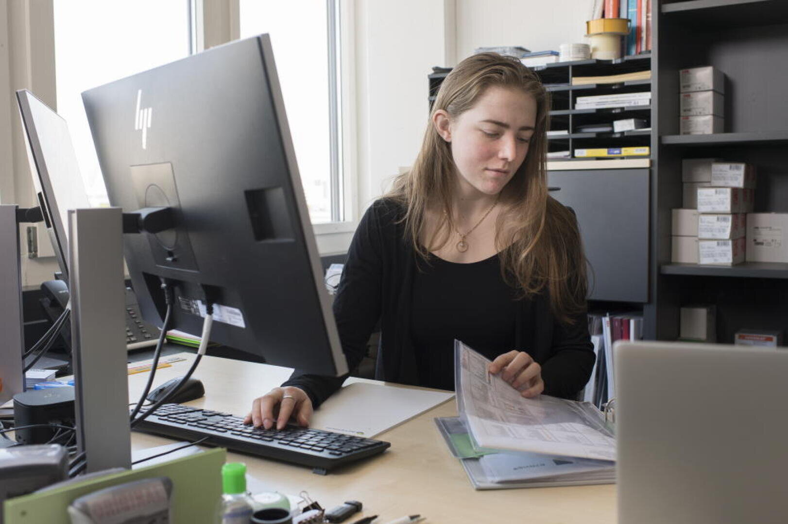 Une femme est assise à un bureau ; d'une main, elle utilise la souris de l'ordinateur, tandis que de l'autre, elle consulte des documents contenus dans un classeur. Elle est entourée de fournitures de bureau, d'un écran, de classeurs et d'étagères sur lesquelles sont posées des boîtes.