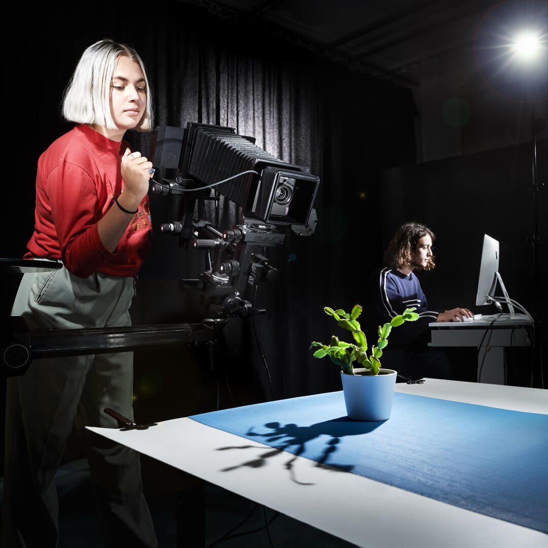 Une femme vêtue d'une chemise rouge manipule une grande caméra braquée sur une plante en pot placée sur une surface bleue, tandis qu'une autre personne travaille sur un ordinateur à l'arrière-plan, sous un éclairage dramatique.