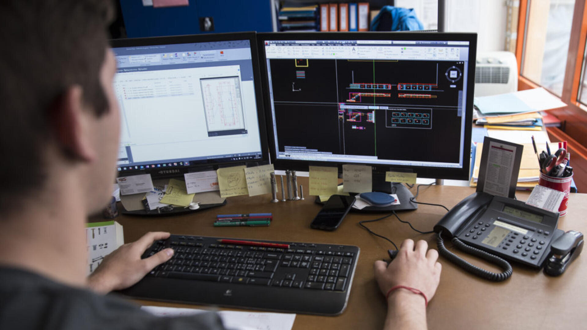 Une personne travaille à un bureau équipé de deux écrans d'ordinateur affichant des dessins et des données techniques. Le bureau est encombré de fournitures de bureau, de papiers, d'un téléphone, de post-its et de stylos.