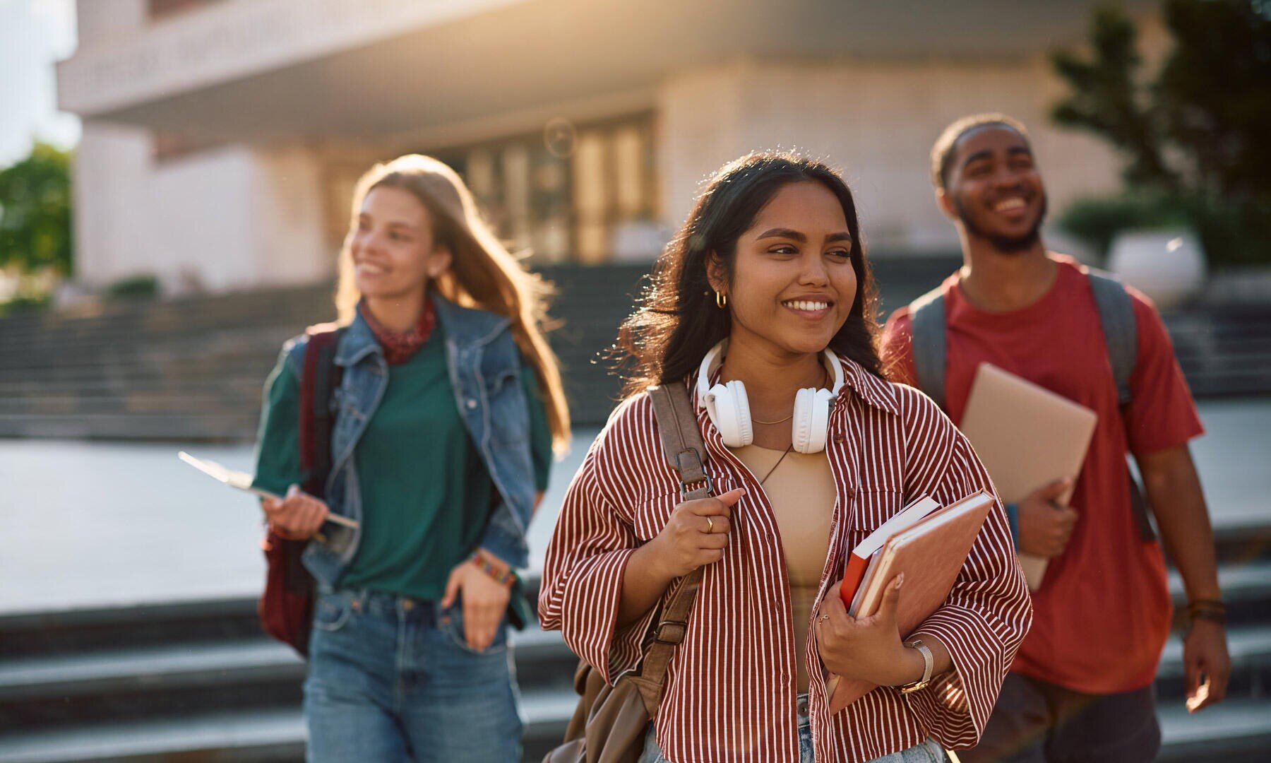 Trois étudiants souriants marchent en plein air sur les marches du campus. L'un d'eux tient des livres et un casque audio, tandis que les autres portent des cahiers. Ils ont l'air heureux et sûrs d'eux, avec la lumière du soleil qui brille derrière eux.