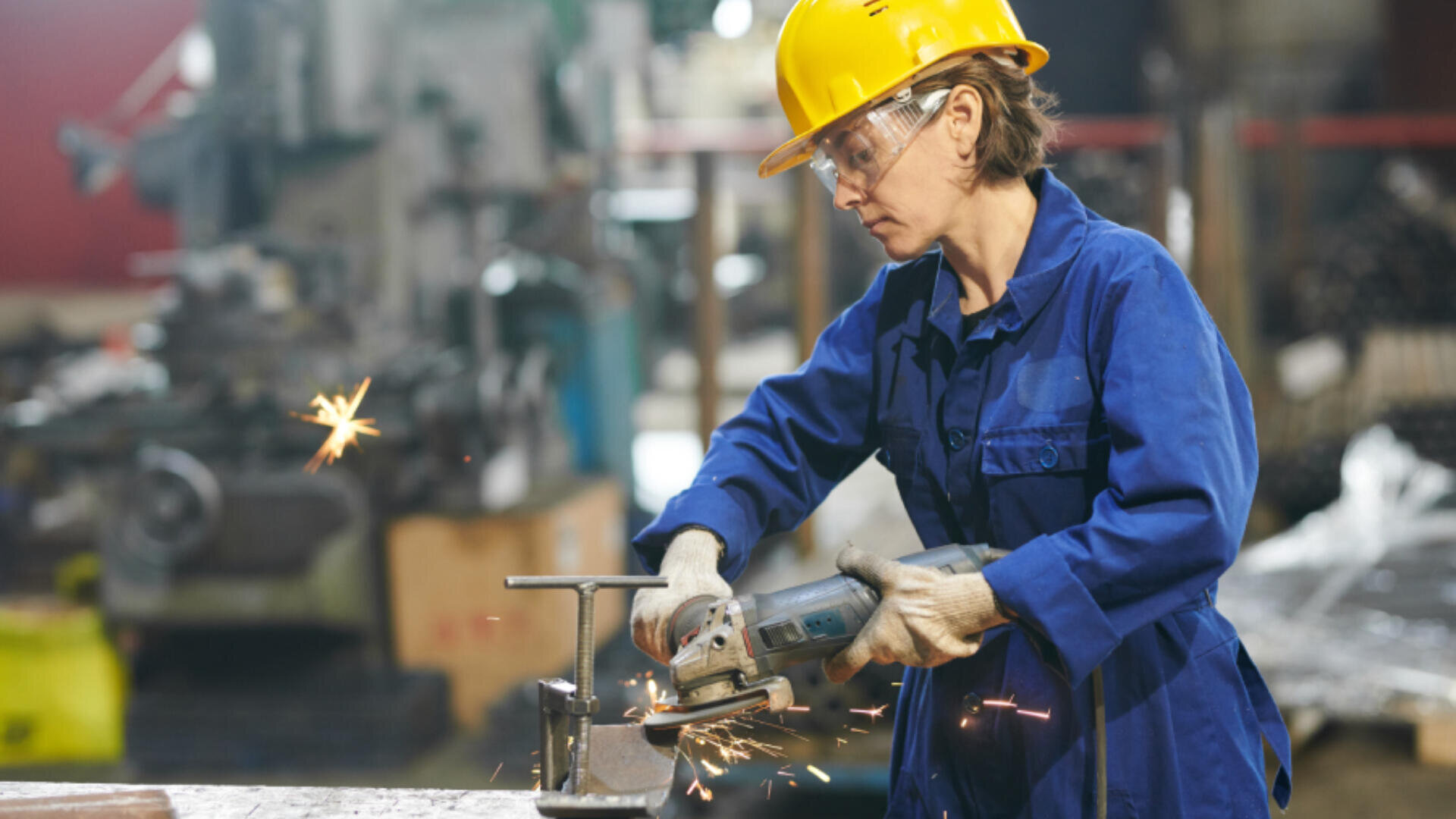 Une personne portant un casque jaune, des lunettes de sécurité, des gants et une tenue de travail bleue utilise une meuleuse d'angle sur du métal, créant des étincelles dans un atelier industriel.