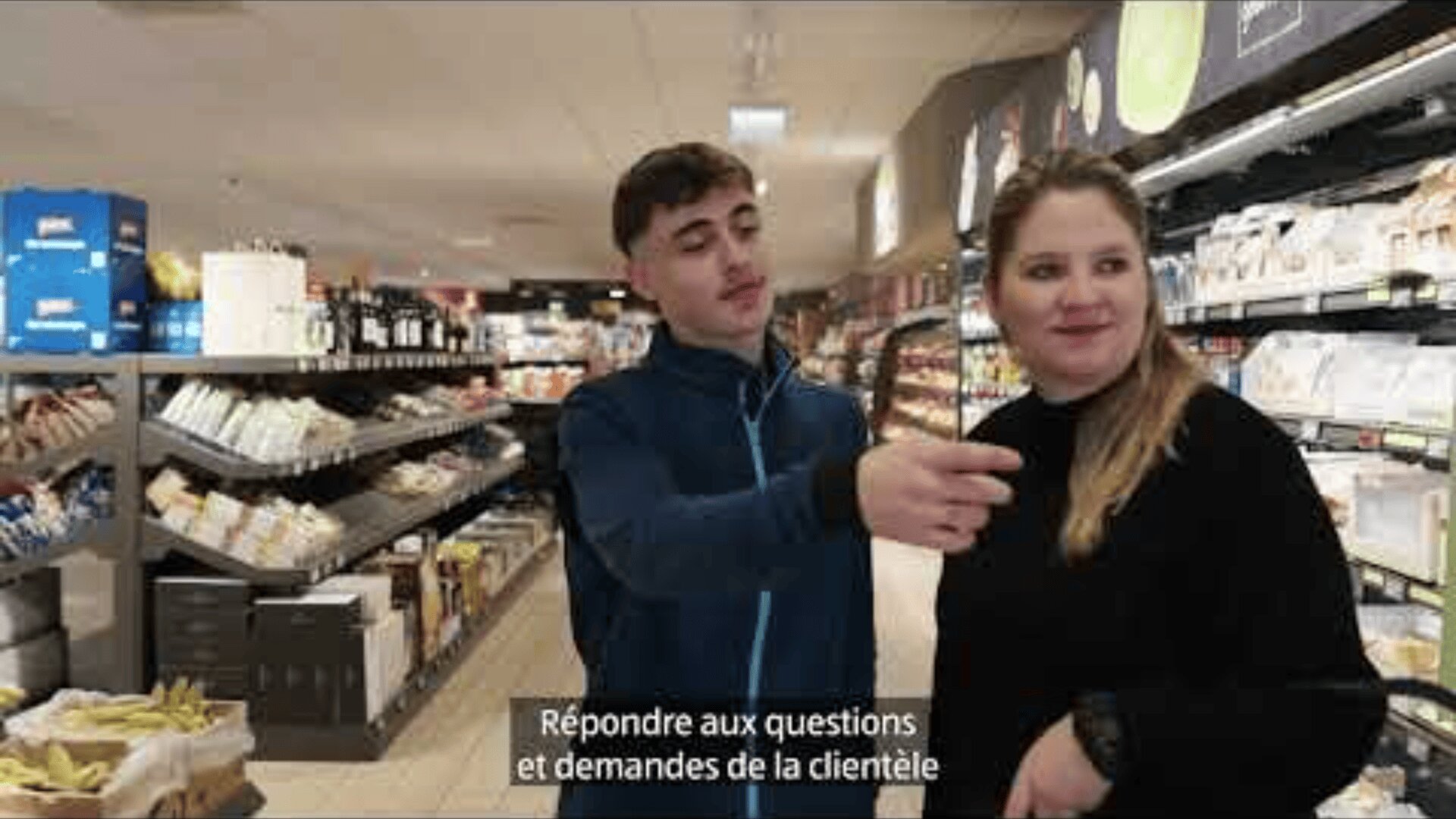 Un jeune homme et une jeune femme se tiennent ensemble dans l'allée d'un magasin d'alimentation. L'homme montre quelque chose du doigt tandis que la femme regarde devant elle en souriant. Les sous-titres en français au bas de la page indiquent : "Répondre aux questions et demandes de la clientèle".
