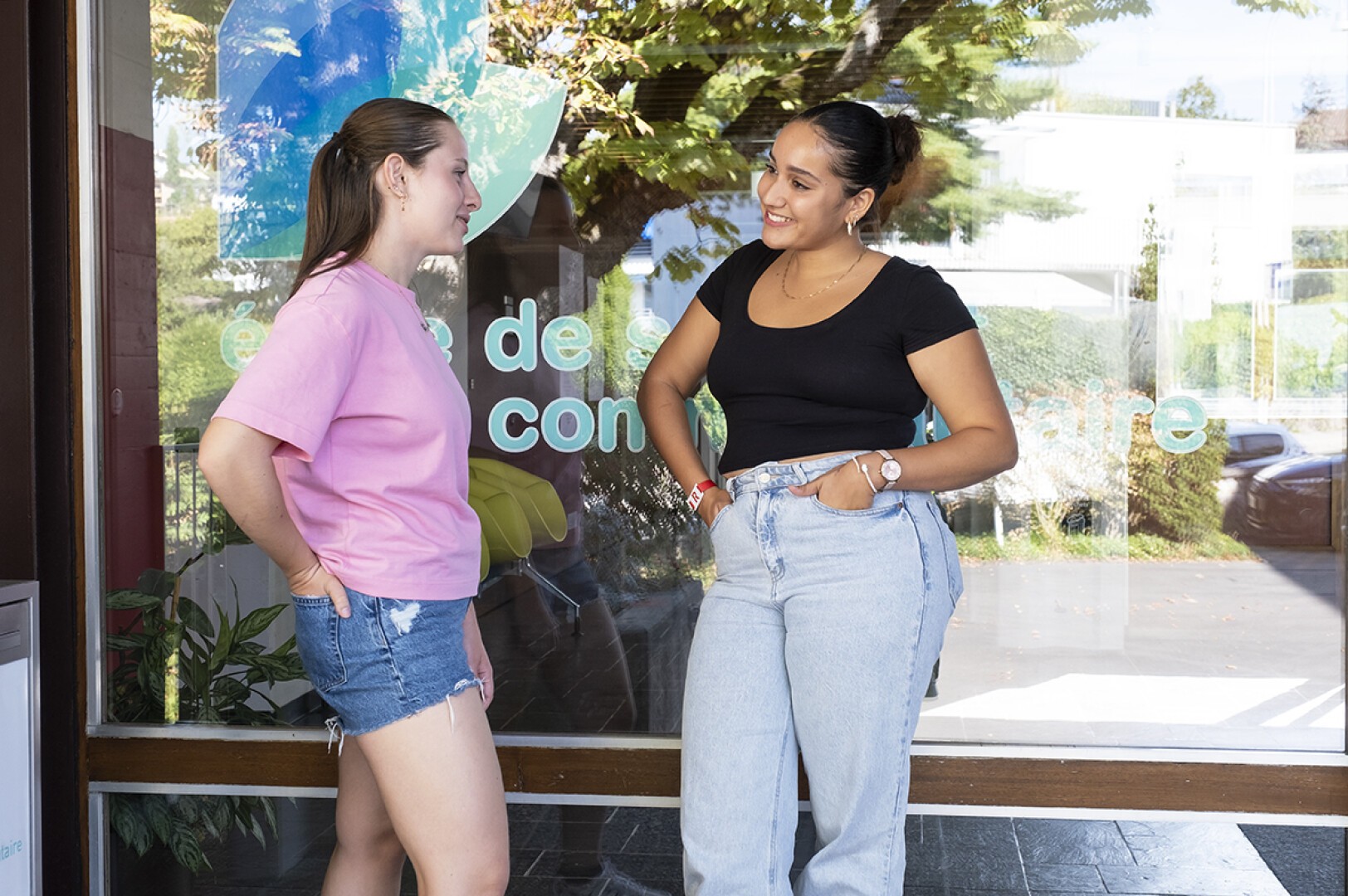 Deux jeunes femmes se tiennent dehors devant une baie vitrée, souriantes et discutant. L'une porte un t-shirt rose et un short en jean, l'autre un t-shirt noir et un jean. Des arbres et des reflets sont visibles en arrière-plan.