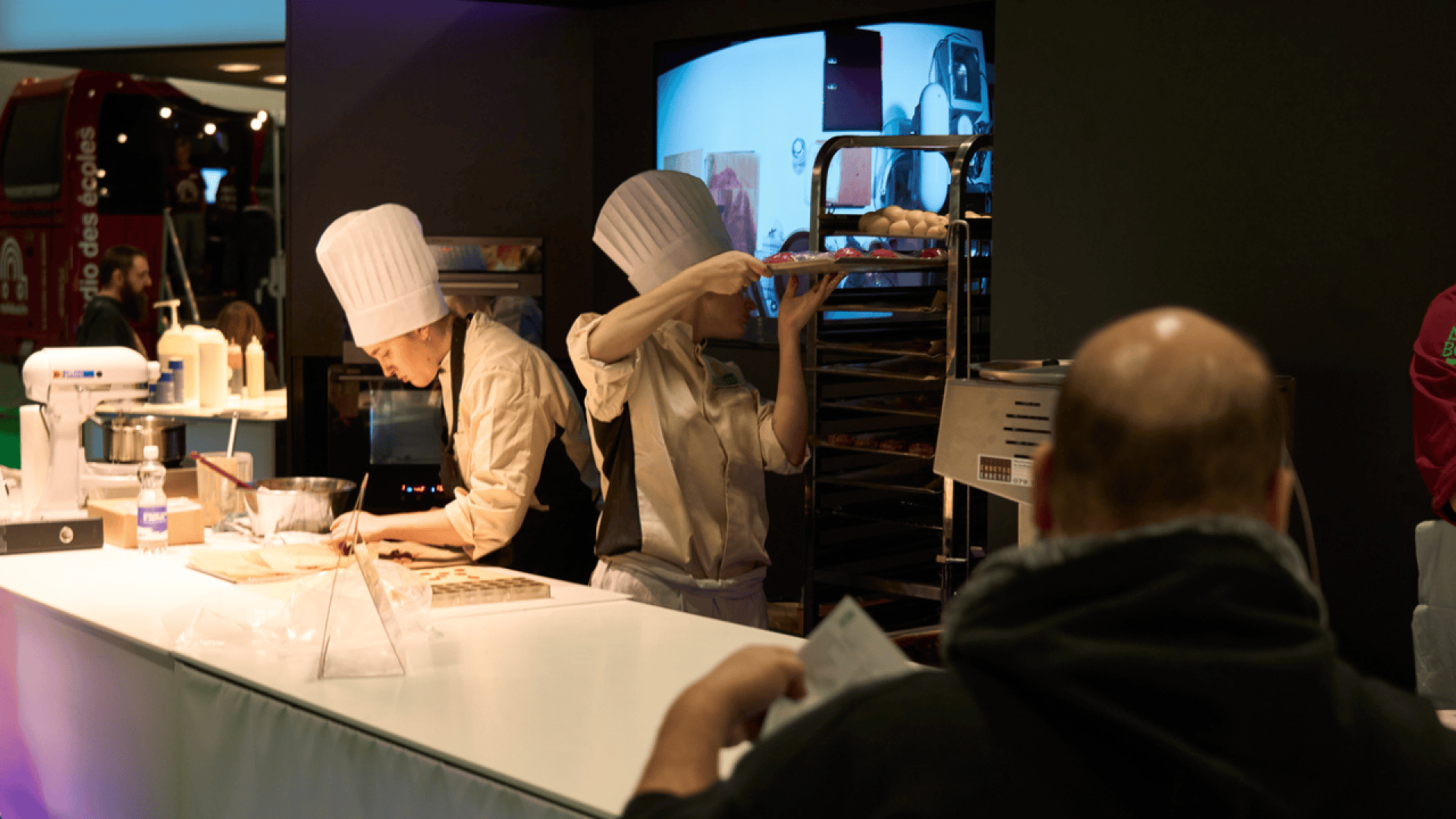 Deux chefs en toque blanche et en uniforme préparent des plats derrière un comptoir. L'un d'eux travaille la pâte tandis que l'autre place des produits de boulangerie sur une grille. Une personne observe au premier plan, partiellement visible.