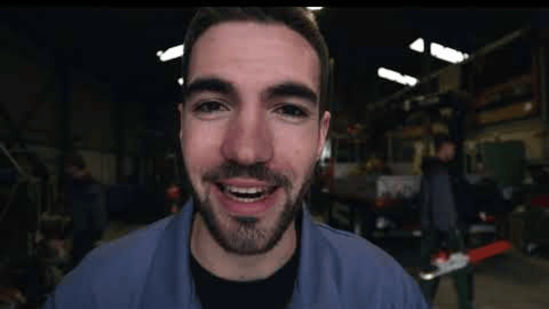 Un jeune homme aux cheveux bruns courts et à la barbe sourit à la caméra dans un atelier industriel, vêtu d'une veste de travail bleue. L'arrière-plan est flou, montrant des équipements et une autre personne en train de travailler.