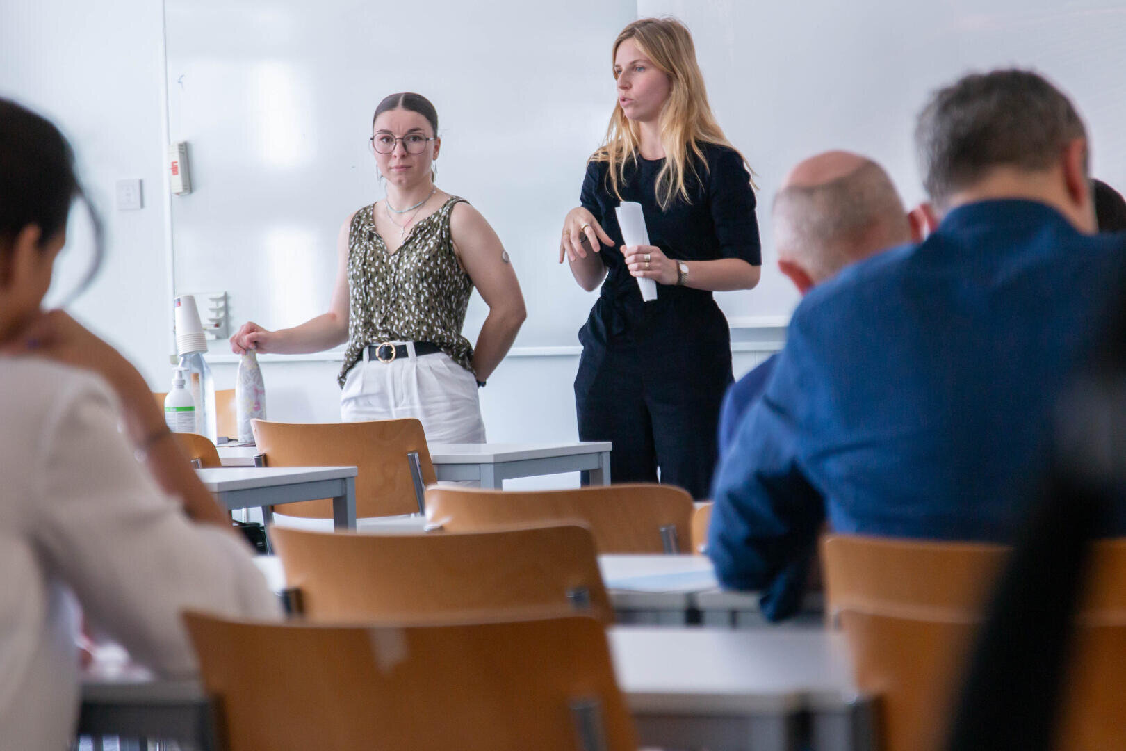 Deux femmes se tiennent à l'avant d'une salle de classe, l'une tenant des documents et parlant, tandis que l'autre écoute. Plusieurs personnes sont assises à des bureaux, face à elles. La scène semble être une présentation ou une conférence.