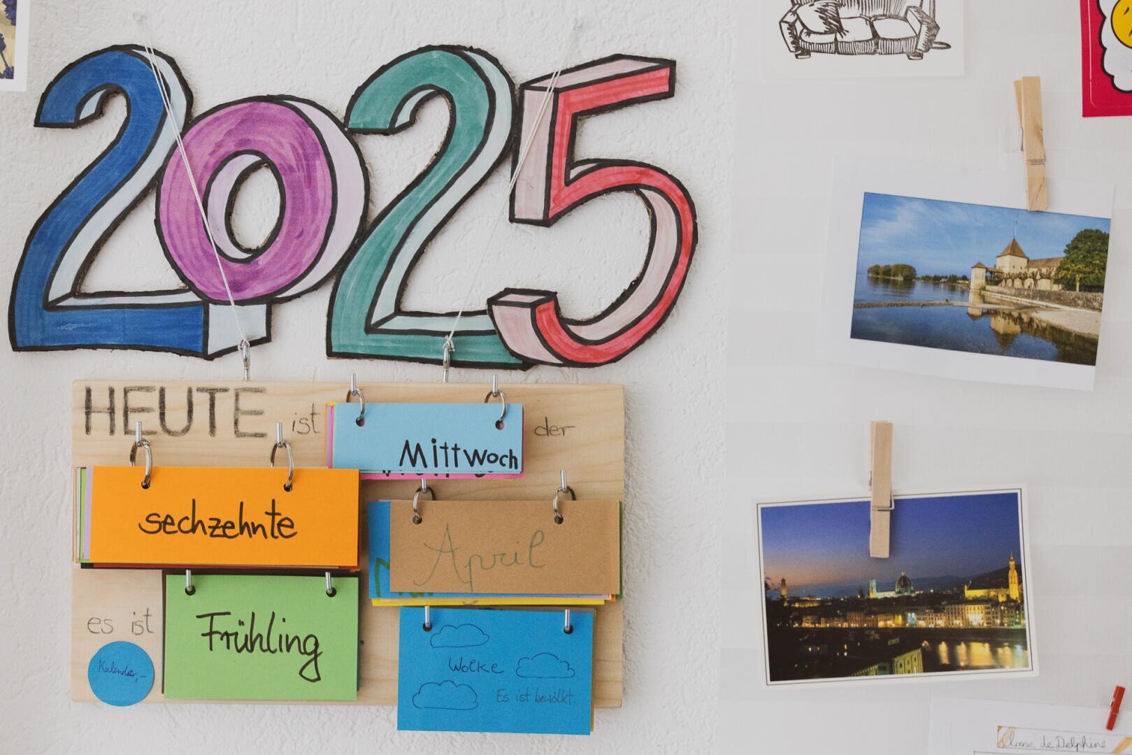 Panneau coloré de 2025 au-dessus d'un tableau affichant des mots allemands pour le jour, la date, le mois, la semaine et la saison avec des cartes manuscrites ; à proximité, deux photos en format paysage sont accrochées au mur blanc.