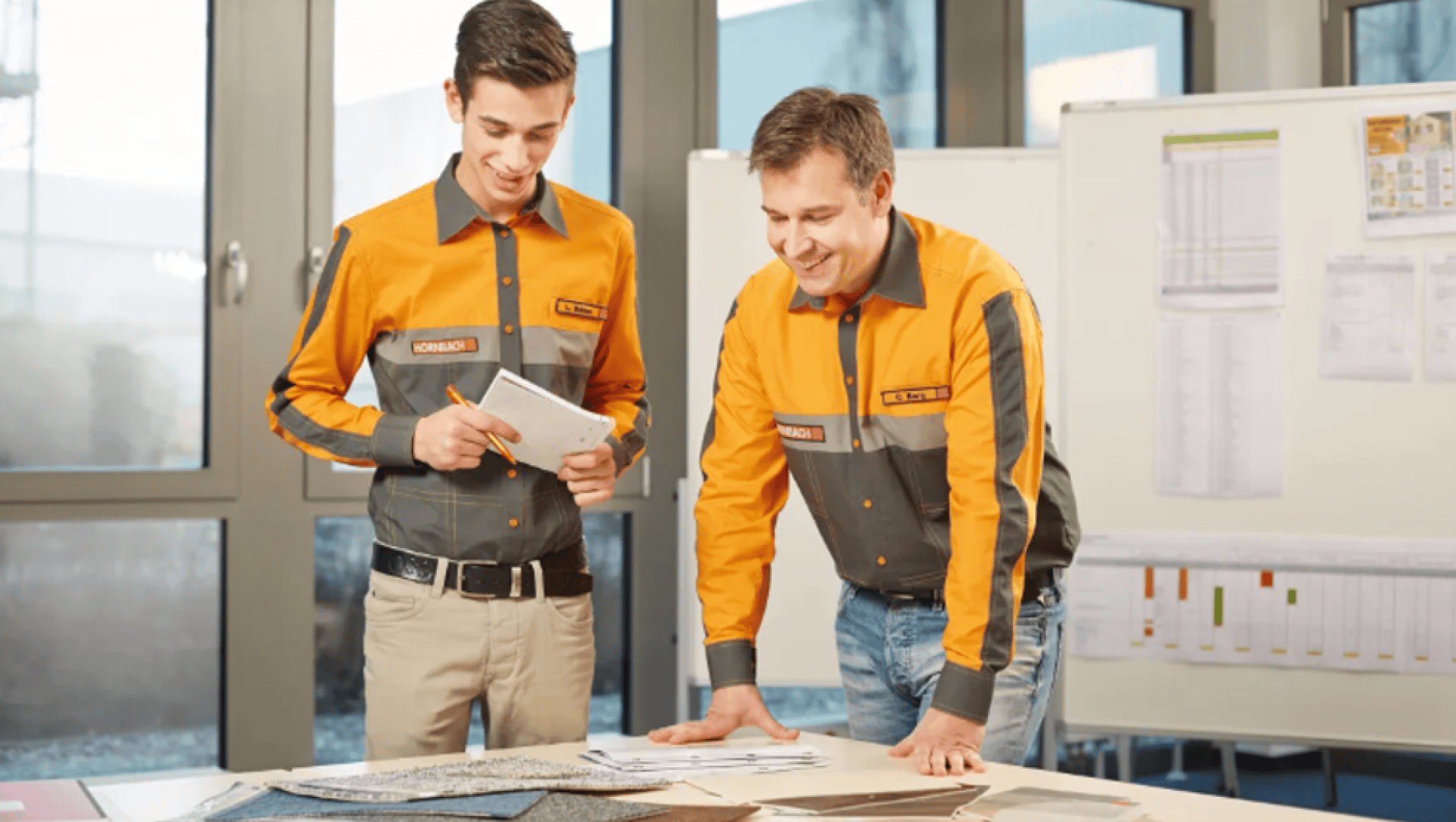 Deux hommes en tenue de travail assortie examinent des documents et des échantillons de matériaux sur une table dans un bureau lumineux, avec des tableaux et des horaires visibles à l'arrière-plan.