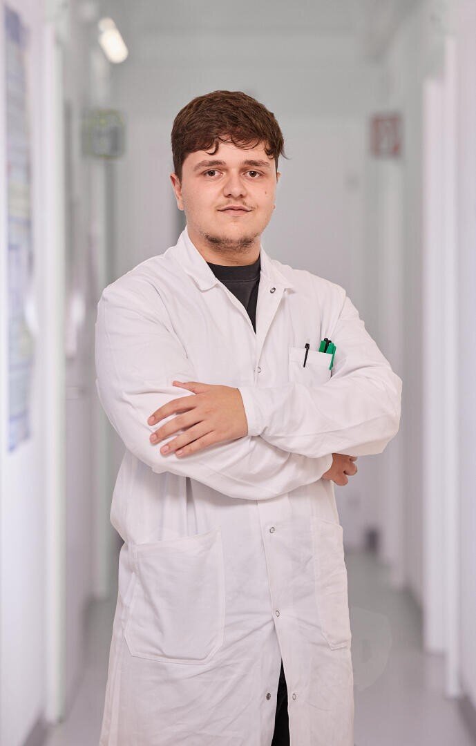 Un jeune homme portant une blouse blanche se tient dans un couloir lumineux, les bras croisés, regardant la caméra avec assurance. Des stylos sont visibles dans la poche de la blouse et l'arrière-plan est flou.