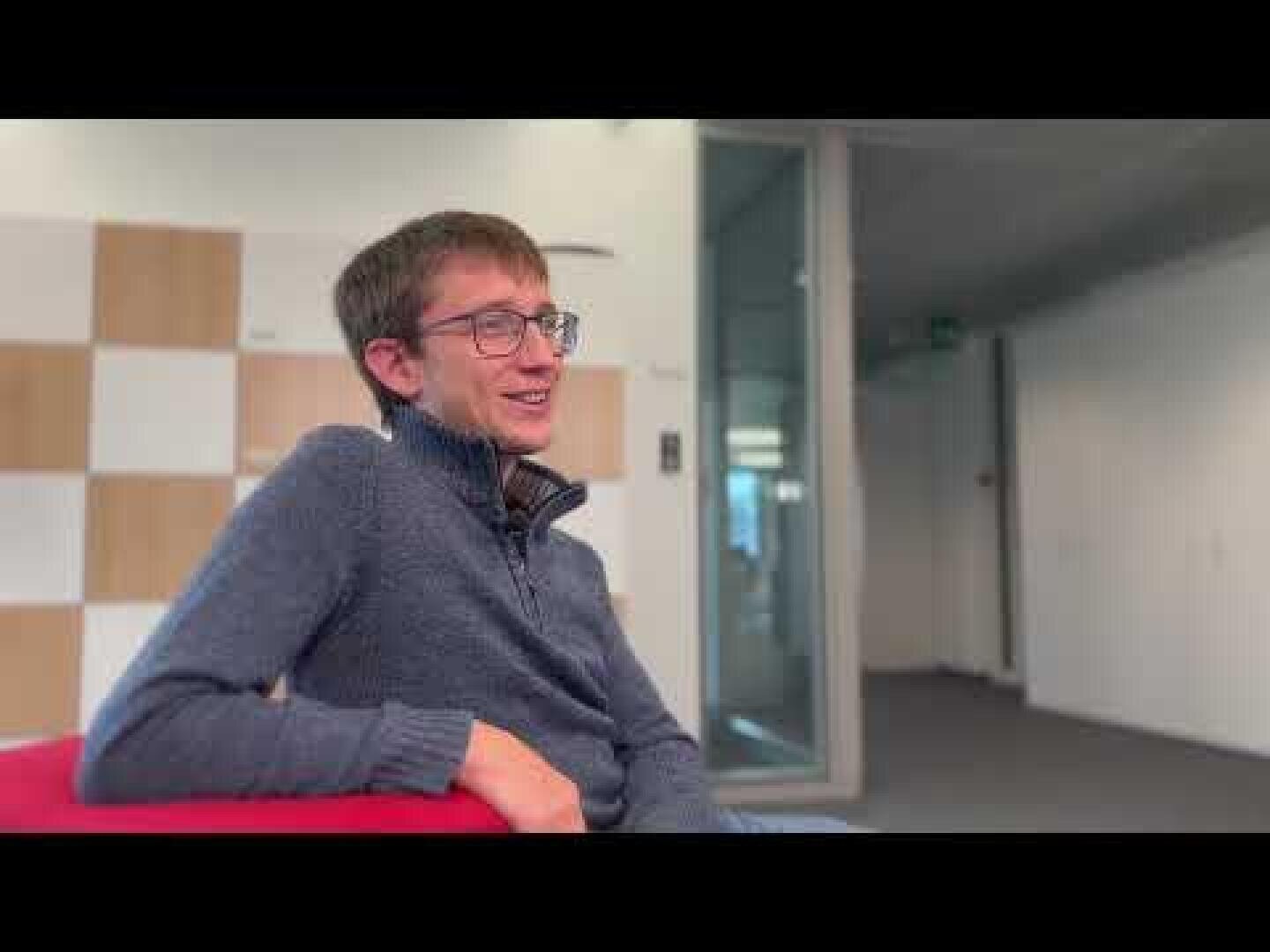 Un jeune homme portant des lunettes et un pull bleu est assis, souriant, dans un bureau moderne avec une porte vitrée et un mur à carreaux en arrière-plan.