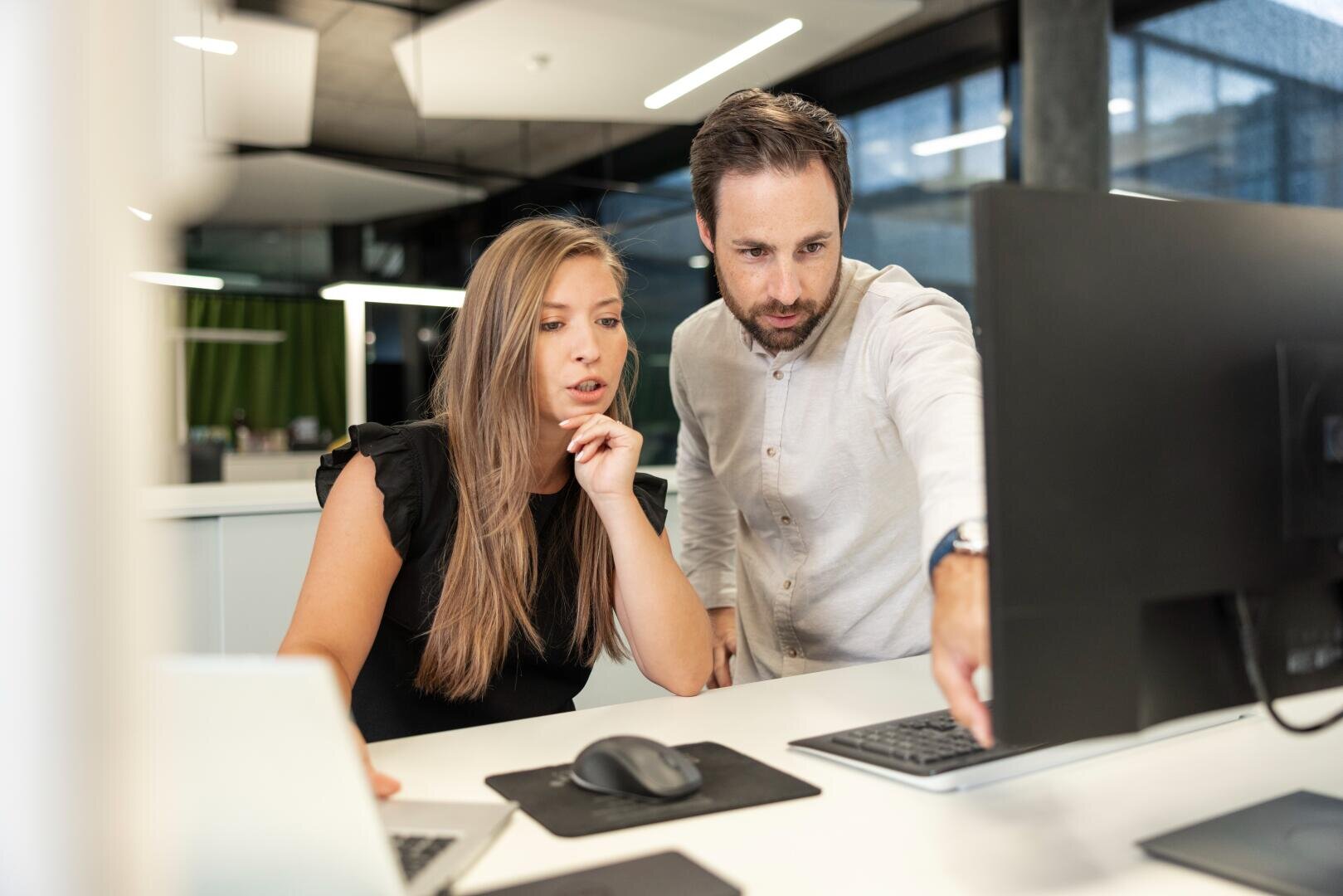 Une femme et un homme travaillent ensemble à un bureau, regardant attentivement un écran d'ordinateur tandis que l'homme pointe du doigt l'écran. L'espace de travail est moderne et éclairé par la lumière naturelle.